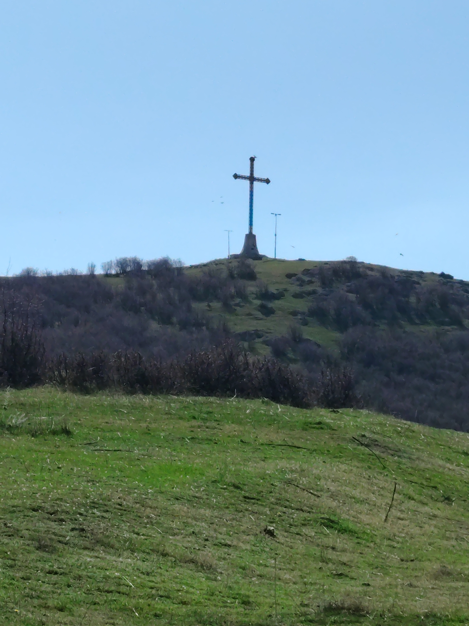 The image depicts a large Christian cross situated on the summit of a hill. The cross is dark in color with what appears to be a blue accent running up the vertical portion of the cross. The hill is covered in green grass with patches of bare earth and bushes. The foreground is also green with sparse vegetation. Two light poles are standing to the right of the cross. The sky is a clear, pale blue. The cross is located in Bata, Bulgaria.