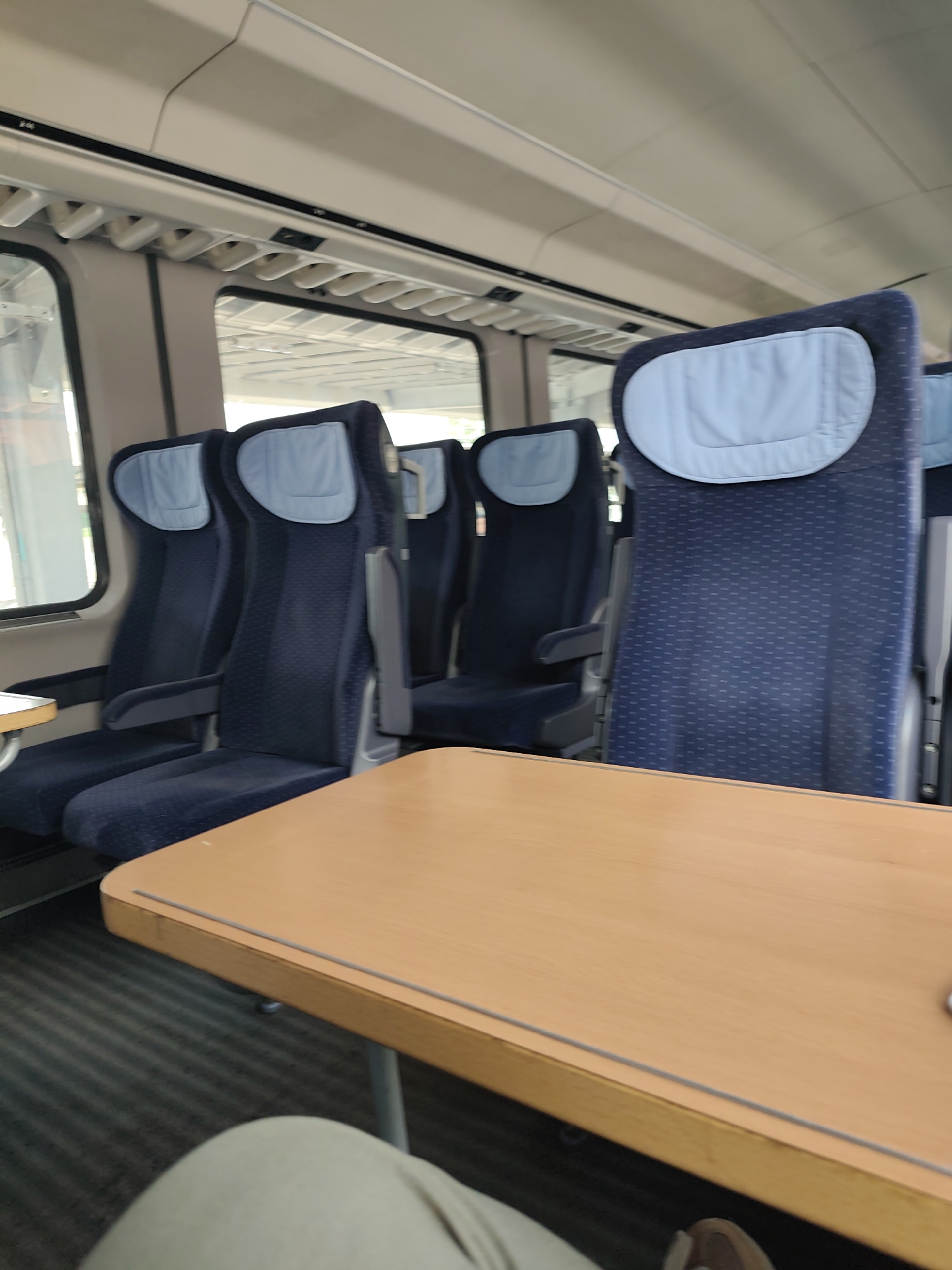 The image shows the interior of a train car, likely a high-speed train given the design of the seats and overhead storage. There are rows of dark blue seats with light blue headrests, and a light brown table is positioned in the foreground, in front of the seats. The train car appears to be empty of passengers, except for a portion of a person's legs and lower torso visible in the bottom left corner, suggesting the photo was taken by someone seated there. The environment is clean and modern, typical of public transportation. The image does not provide any visual cues that would associate it with the city of Plovdiv, Bulgaria.