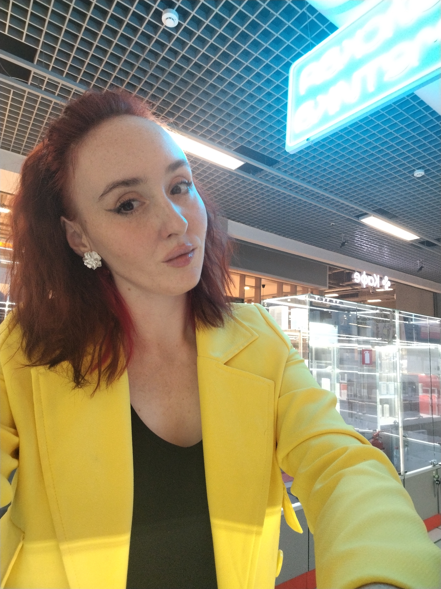 A woman with fair skin, red hair, and freckles is posing for a selfie in what appears to be a commercial or retail space. She is wearing a bright yellow blazer over a dark top and has floral earrings. In the background, there is a ceiling with a grid-like pattern and a glowing blue sign. Further in the background, a cafe is visible, with a sign in Russian, "Кафе". Based on this information, it's plausible this photo was taken in a shopping center or similar commercial area in the city of Izhevsk, Russia. The woman is holding the camera, and the perspective suggests a first-person view.