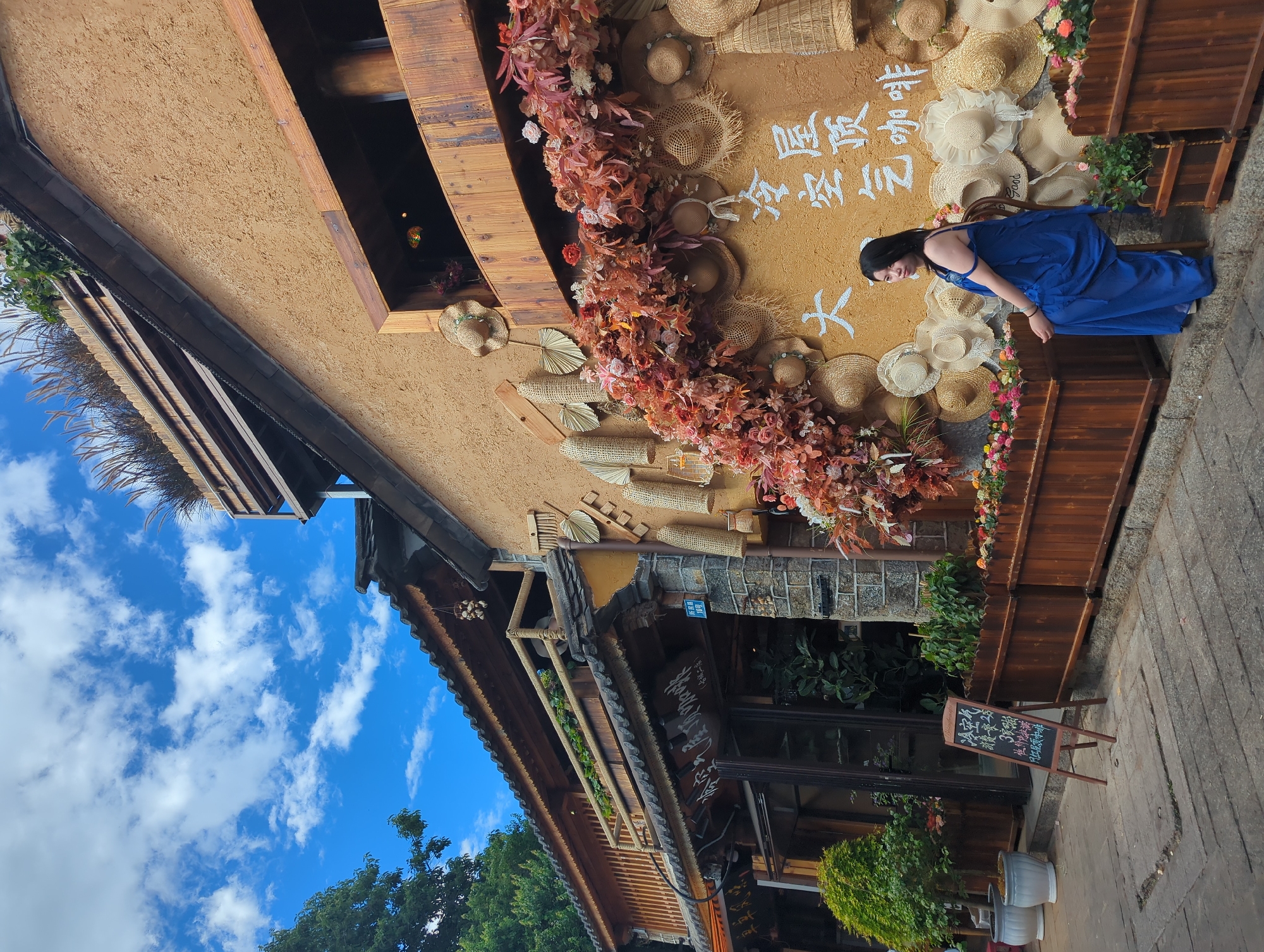 A woman in a blue dress is leaning against a textured wall decorated with straw hats and autumn-colored flowers. She is looking down, and her posture suggests a relaxed or contemplative mood. The wall also features white Chinese calligraphy. To the left of the wall, a traditional building with a thatched roof and wooden accents is visible, with a bright blue sky dotted with white clouds above it. The setting appears to be outdoors, possibly in a marketplace or along a street in Dali City, China, given the architectural style and the Chinese text. A wooden planter box filled with greenery and flowers is situated in front of the wall. A black chalkboard sign with colorful writing stands on the pavement in front of the planter box. The overall scene evokes a sense of calm and perhaps a tourist destination. The visible text mentions "Cold Air Rooftop Coffee" and "Good value for money," suggesting the location might be a cafe or a shop. The time of day appears to be daytime, with bright sunlight indicated by the shadows and the clear sky.