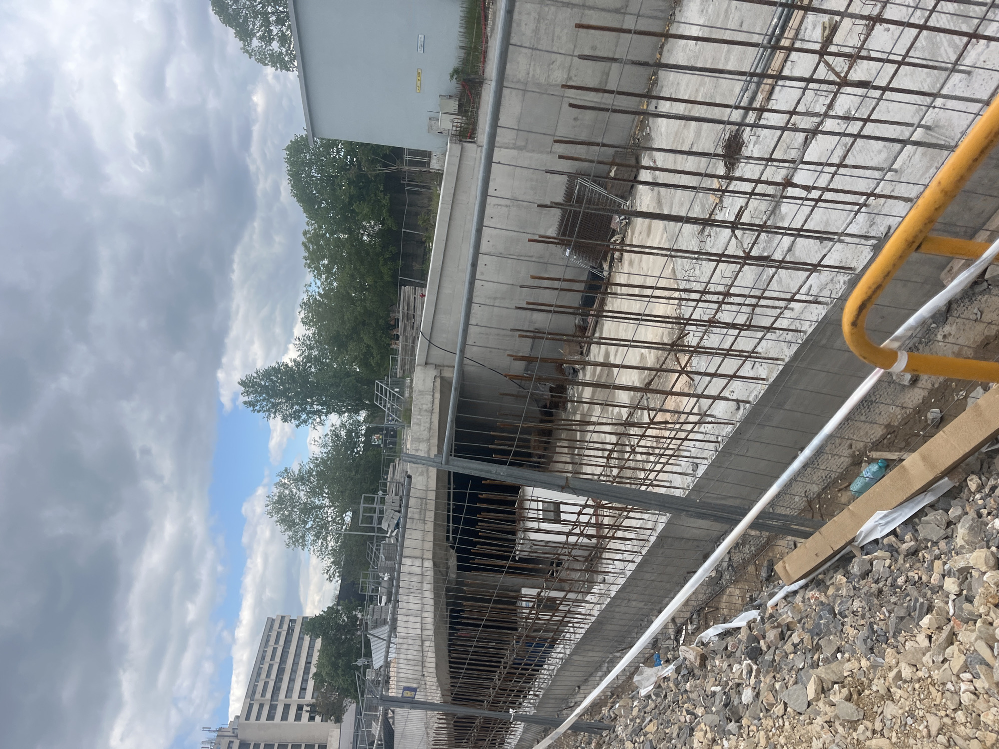 The image depicts a construction site, likely in Пловдив, Bulgaria, given the cityscape visible in the background. A concrete structure, possibly a bridge or building foundation, is under construction. Rebar reinforcement is prominent throughout the scene. A temporary safety fence with yellow and grey components surrounds the construction area. The ground is covered with gravel. In the background, buildings and trees suggest an urban environment under cloudy skies. No people are visible in the frame.