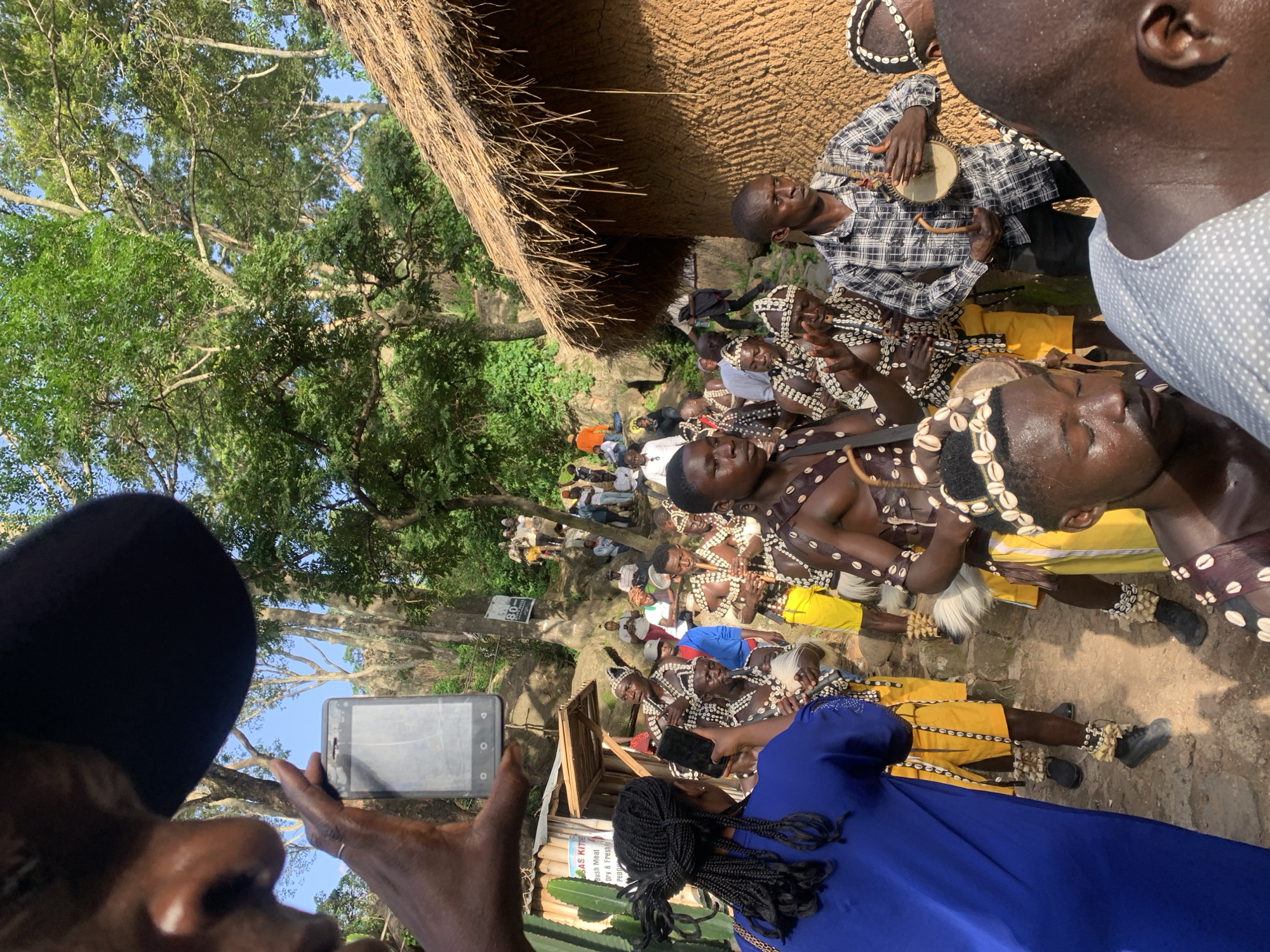 This media file captures a gathering of people in an outdoor setting, likely Jos, Nigeria. The scene appears to be a cultural event or performance, with several individuals adorned in traditional attire featuring shell necklaces and bracelets, and some wearing yellow skirts or sashes. One person in the foreground is playing a drum, while others seem to be observing or participating in the event.
The backdrop includes a rustic structure with a thatched roof and mud walls, suggesting a traditional village or compound. Lush green trees and vegetation surround the area, indicating a daytime scene under clear skies. The presence of a smartphone in the foreground, held by a person, suggests a modern element integrated with the traditional elements.
The subjects are primarily men, with one individual in the foreground wearing a blue dress. Their expressions and the overall atmosphere suggest engagement and celebration. The sunlight filtering through the trees points to a sunny day, likely mid-morning or afternoon. There are no clearly legible text elements visible in the image.