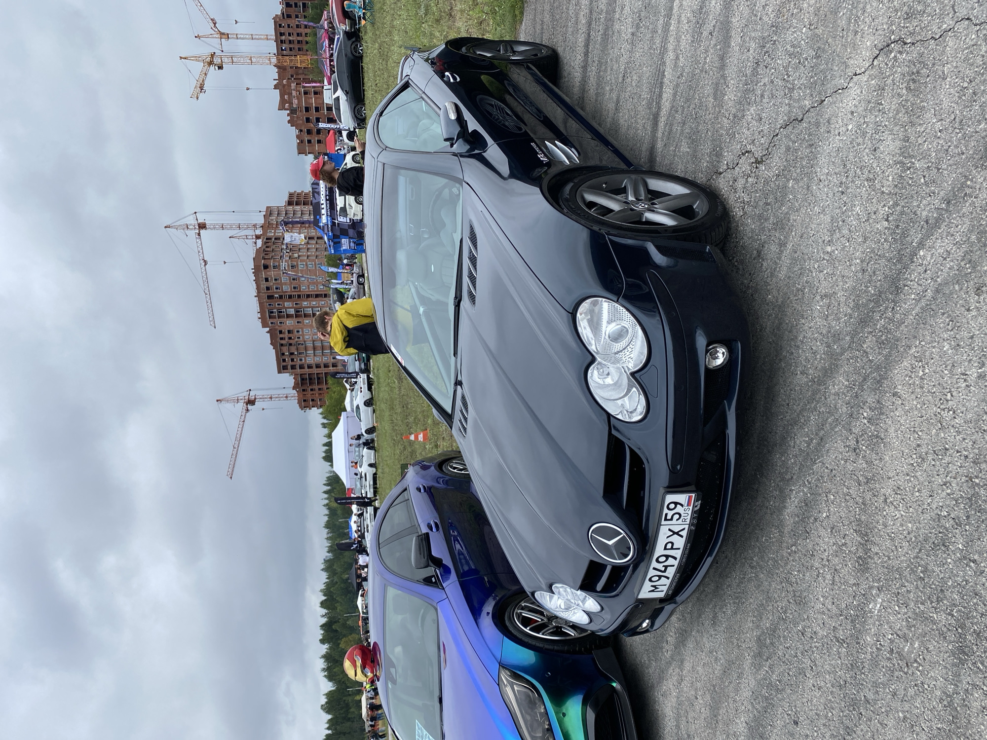 In Perm, Russia, on an overcast day, two cars are parked on an asphalt lot. In the foreground, a dark blue Mercedes-Benz SL-Class convertible is parked, its front license plate reading "M949 PX 59 RUS". To the left of it, a blue and green iridescent car is parked, with a red and gold helmet on its roof. Behind the cars, a crowd of people are gathered, with tents and construction cranes visible in the background. The sky is cloudy.