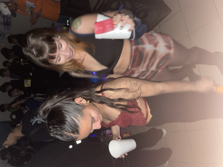 The image depicts an indoor social gathering, likely a party or club, in Ushuaia, Argentina, under dim lighting. Two women are prominently featured in the foreground, with numerous other individuals blurred in the background.
The woman on the left has dark hair pulled into a high ponytail. She wears gold-colored hoop earrings and an ear cuff. Her attire consists of a dark red, lace-textured bralette exposing her midriff, paired with dark pants. She holds a white disposable cup in her left hand, with her head tilted downwards.
The woman on the right has lighter brown hair with bangs. She wears a black v-neck crop top, a short skirt with a red, black, and white abstract pattern, and dark tights. A green and yellow tattoo is visible on her left upper arm. In her right hand, she holds a white disposable cup containing a red popsicle or ice bar. A dark blue strap, possibly from a bag, is visible on her left shoulder. Her facial expression indicates a smile, directed downwards and towards the woman on the left.
The background shows a crowded, out-of-focus scene of people standing and socializing. One individual on the far right wears an orange shirt and a clear face shield, appearing to interact with a smartphone. The floor is light-colored tile. A dark speaker is partially visible on the right. The overall environment is dimly lit with ambient blue and purple lighting contributing to the atmosphere.