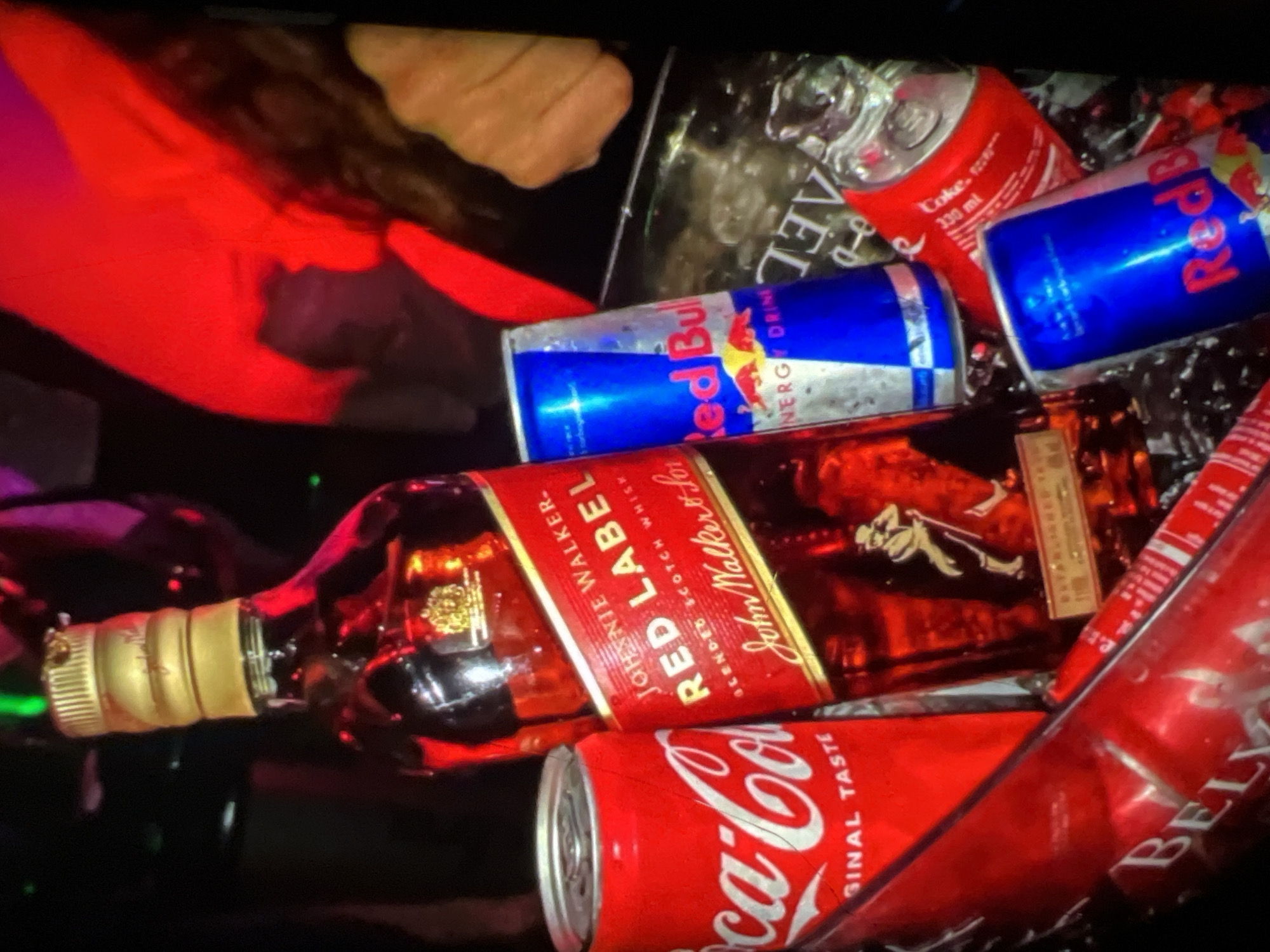 The image depicts a collection of alcoholic and non-alcoholic beverages, including a bottle of Johnnie Walker Red Label whiskey, several cans of Coca-Cola, and at least two cans of Red Bull. The scene appears to be in a dimly lit environment, possibly a bar or a private party, with red and purple lighting casting a glow on the objects. One of the Coca-Cola cans has text that includes "GINAL TASTE". The Johnnie Walker bottle features the iconic Striding Man logo and text that reads "RED LABEL BLENDED SCOTCH WHISKY". One of the Red Bull cans is partially visible and shows its distinct blue and red branding with the Red Bull logo. There is also a glimpse of a person's hand in the background, suggesting a social gathering. The overall impression is that of a celebratory or social occasion where drinks are being consumed. The image does not contain any specific visual cues that would allow for its precise location to be identified as Lagos, Nigeria.
