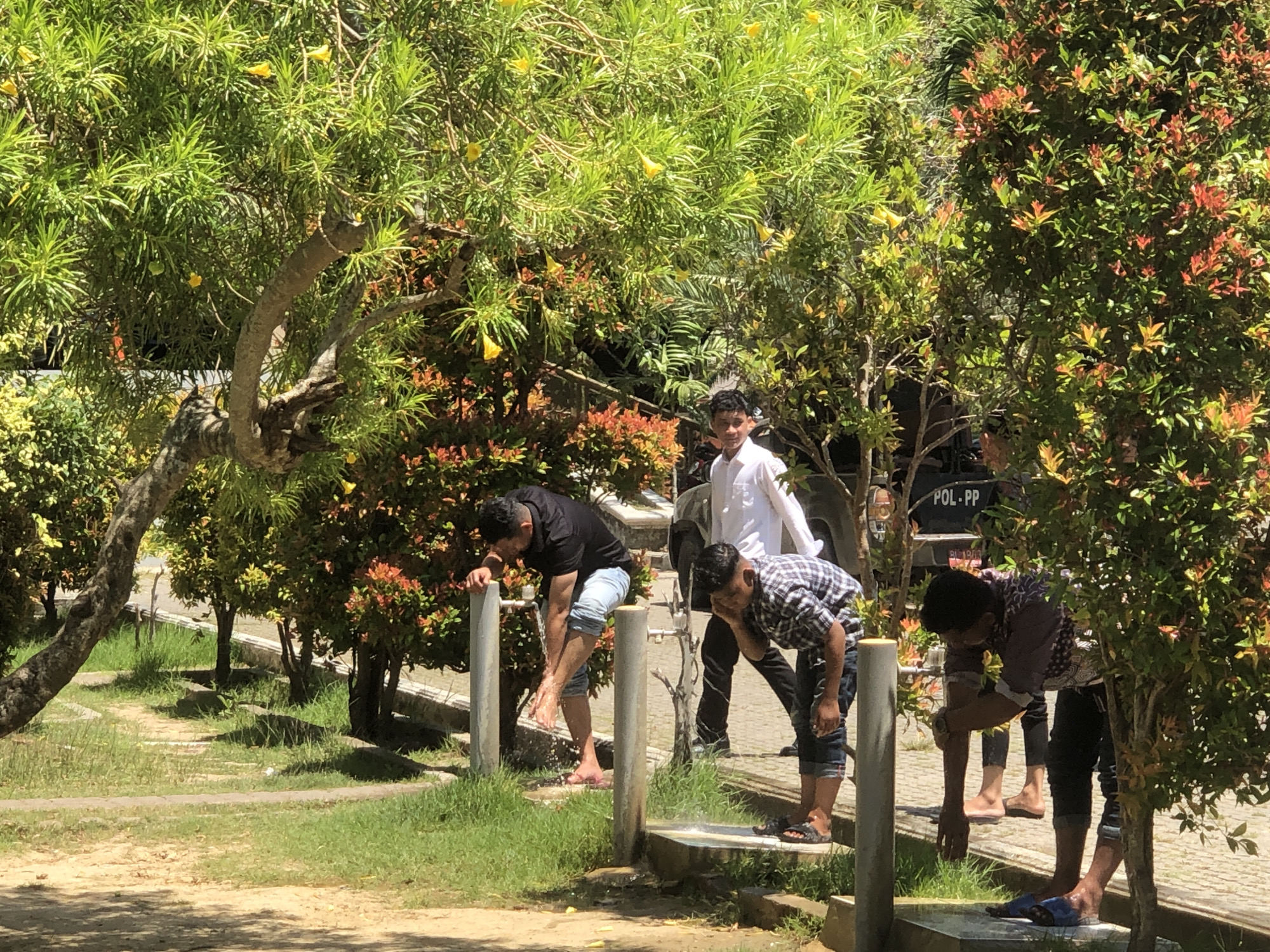 The scene is an outdoor ablution area in the city of Lhokseumawe, Indonesia. Four men are performing ritual washing before prayer. Each stands at a concrete pillar equipped with a water faucet, washing their hands, feet, and faces. The man farthest to the left is wearing a black t-shirt and light-colored shorts. The man in the center is wearing a checkered shirt and dark shorts. The man on the right wears a patterned shirt and dark pants. A fourth man, dressed in a white shirt, stands further behind them. Behind the ablution area, there is a vehicle with "POL - PP" on its side, partially obscured by trees. The area is surrounded by greenery, including trees and shrubs. The ground is paved with light-colored stones.