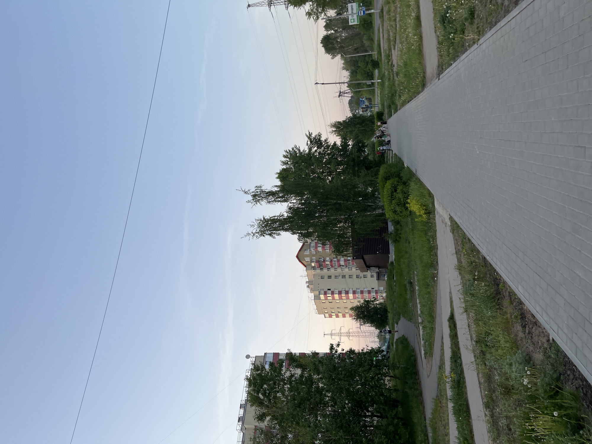The image shows a wide, paved sidewalk running into the distance in the city of Chaikovsky, Russia. A narrower paved path parallels it on the left, separated by a strip of grass. Several people are seen walking together on the sidewalk. Apartment buildings, partly obscured by trees, are visible in the background. A power line runs across the upper part of the image, and transmission towers are present near the center. The sky is a light blue, suggesting early evening.