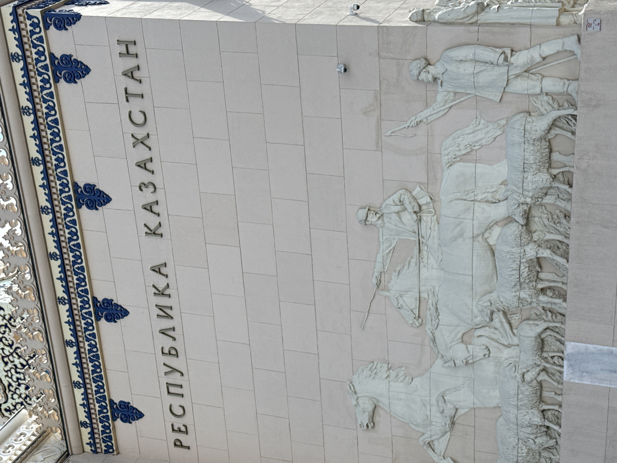 The image shows a relief sculpture on the facade of the Kazakhstan Pavilion at VDNKh (Exhibition of Achievements of National Economy) in Moscow, Russia. The relief depicts a pastoral scene, with two horses and a herd of sheep. Two men are shown on horseback, one appearing to be a shepherd with a staff, and another man, also on horseback, is depicted in the background. The inscription on the facade above the relief reads "РЕСПУБЛИКА КАЗАХСТАН" (Republic of Kazakhstan) in Cyrillic letters. The relief is made of pale stone and is set against a background of similarly colored stone blocks. Decorative blue and gold elements are visible at the top of the facade. Two security cameras are mounted on the wall.