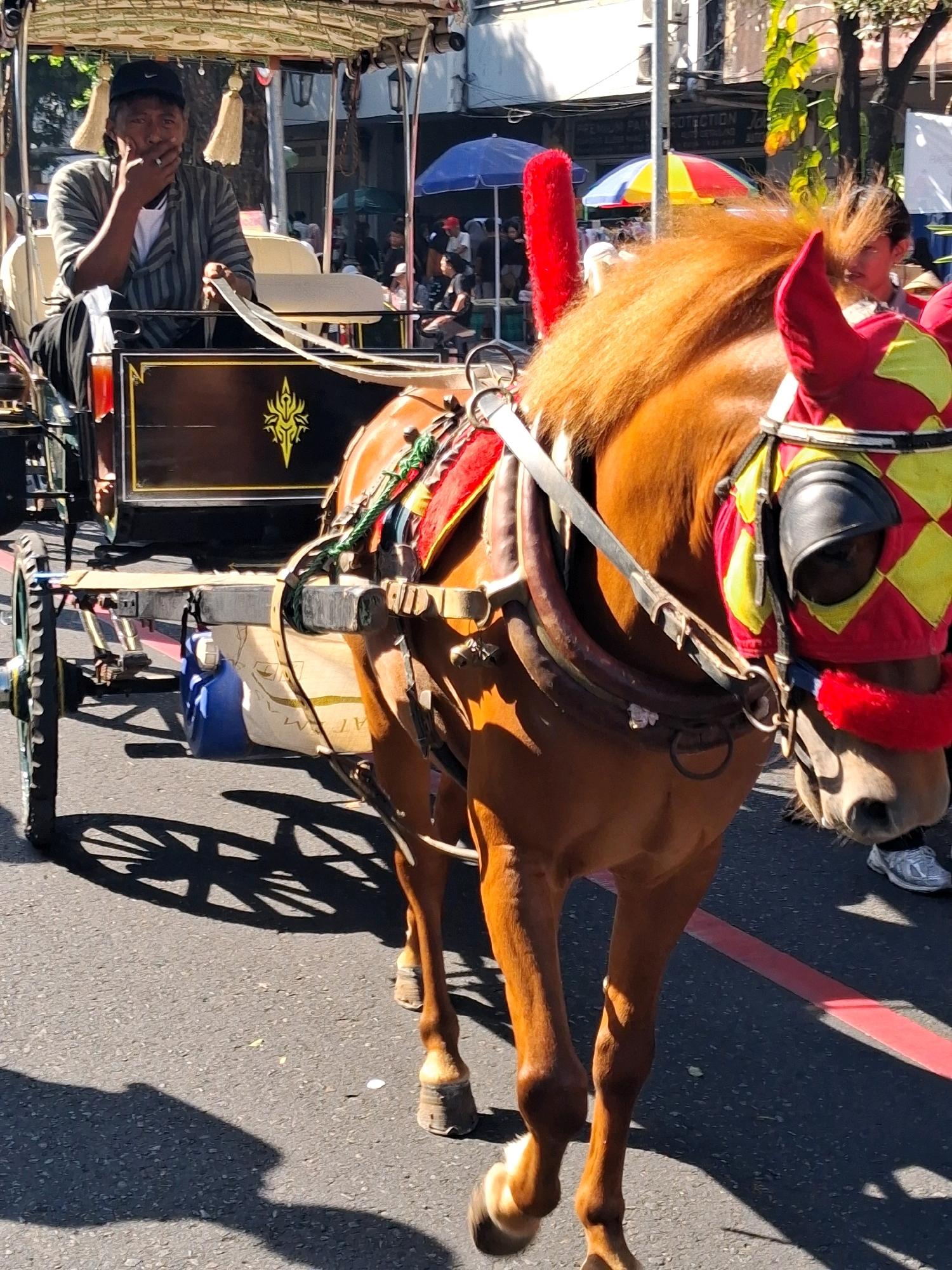 A horse-drawn carriage, known as a *andong*, is in motion on a paved road in Surakarta, Indonesia. The *andong* is decorated with red and yellow checkered fabric and red tassels. A brown horse is harnessed to the front of the carriage, wearing decorative coverings over its eyes. The carriage itself is dark brown with gold trim. A driver, wearing a black cap and a striped shirt, sits in the front of the carriage, holding the reins and smoking. In the background, a colorful umbrella and other pedestrians are visible. The scene suggests a sunny day, with shadows cast by the carriage and horse.