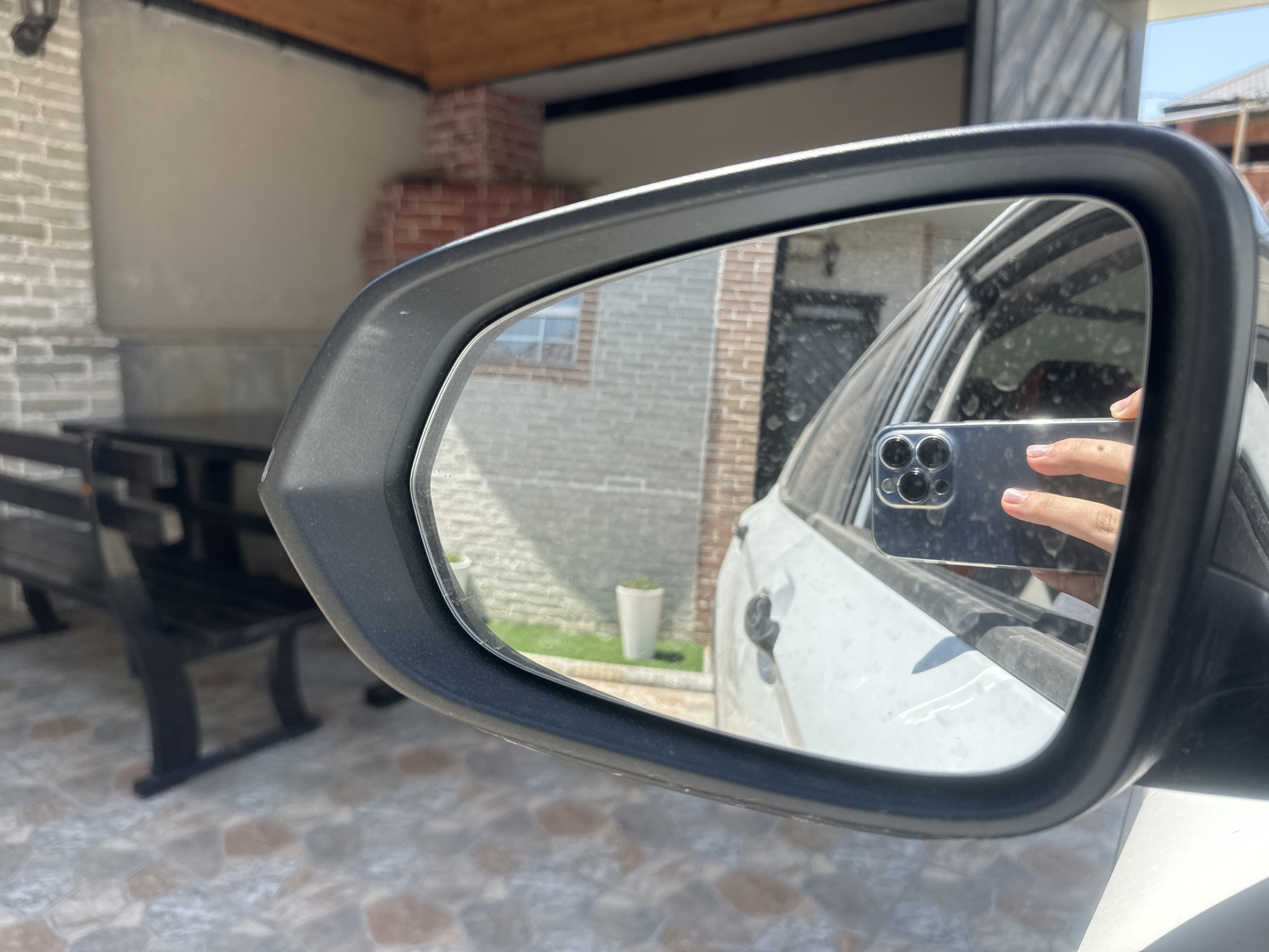 The image is a close-up of a car's side-view mirror, reflecting a person's hand holding a smartphone. The background visible in the mirror shows a building with a brick facade and a white potted plant on a patch of grass. Outside the mirror, a black bench and table are visible on a patterned tiled surface, and a brick wall and wooden ceiling are also partially visible. The scene appears to be in an outdoor area, possibly a courtyard or patio. The car appears to be white. The location is the city of Masazir, Azerbaijan.