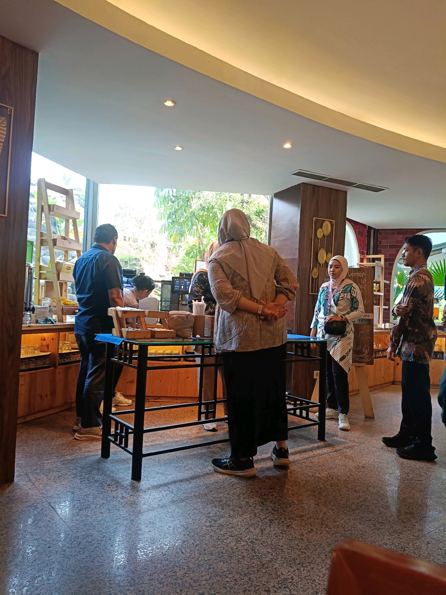 The image depicts an indoor scene, likely a cafe or restaurant, with an open-concept design. The setting appears to be in North Jakarta, Indonesia, suggested by the presence of decorative elements and the general ambiance.
In the foreground, several people are present. On the left, a man in a dark blue shirt and black pants stands facing away from the camera, looking towards a counter area. Next to him, a woman in a light-colored hijab and patterned clothing is partially visible. In the center of the image, a woman wearing a beige hijab and a dark skirt is standing with her hands clasped behind her back. To her right, another woman in a patterned hijab and long-sleeved tunic is interacting with someone at the counter. On the far right, a man in a patterned shirt and dark pants stands facing the other individuals.
The environment features wooden paneling on the walls and counter area, with shelves displaying various items. A large window on the left provides a view of lush green trees and foliage, suggesting an outdoor space beyond. Natural light streams in, illuminating the speckled floor and creating reflections. A black metal table with a wooden top is positioned in the middle of the scene. In the background, there are more shelves, a coffee machine, and decorative artwork on the wall. The overall atmosphere suggests a casual gathering or a service area within a establishment.
