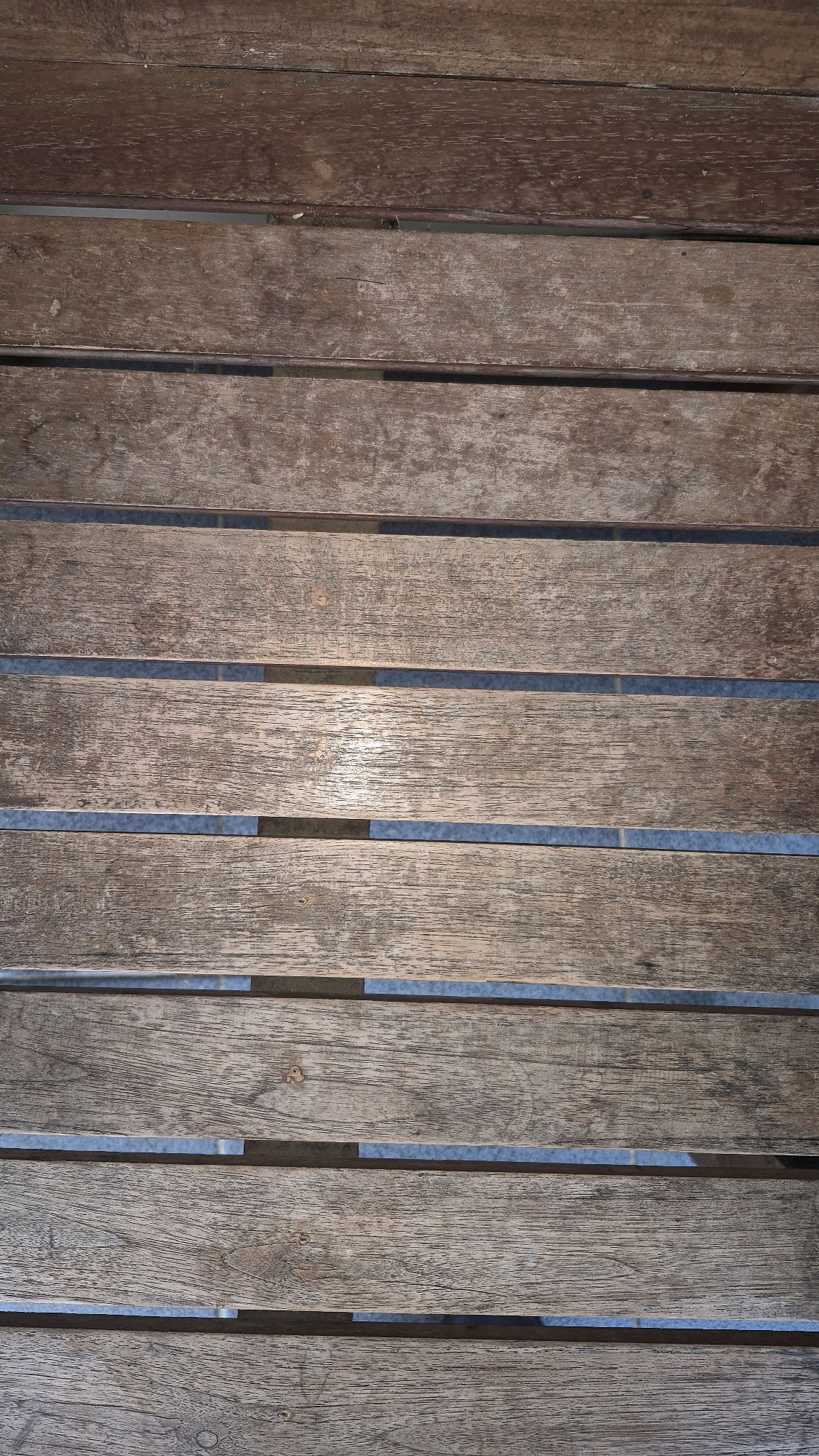The image is a close-up, vertical shot of weathered wooden planks, possibly part of a deck or table. The planks are oriented horizontally and are spaced slightly apart, revealing a blue-gray material beneath them. The wood shows signs of age and exposure to the elements, with variations in color and texture, including some splintering and wear. Sunlight reflects off the surface of some planks, creating highlights. The scene is devoid of people or specific identifiable landmarks, making it impossible to ascertain a specific location within Sukoharjo, Indonesia.