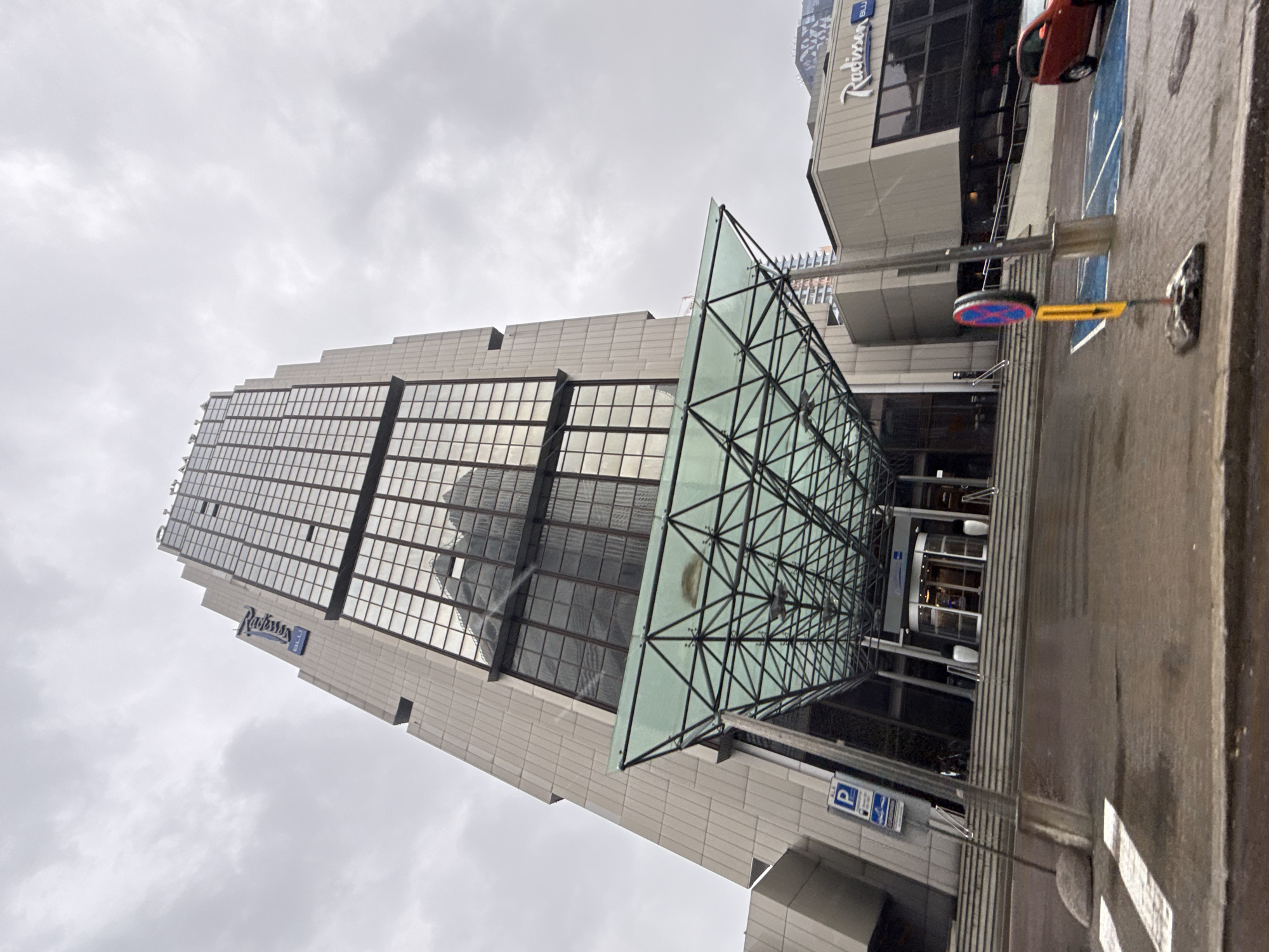 The image shows the Radisson Blu Hotel Tallinn, a skyscraper located in Tallinn, Estonia. The building has a modern architectural style with a glass facade reflecting the overcast sky. The entrance features a prominent glass canopy supported by a metal framework. A revolving door is visible beneath the canopy, along with a set of stairs leading up to it. To the left of the entrance, there is a parking sign and a pedestrian crossing. In the foreground, the wet pavement suggests recent rain. A red car is parked in a blue-marked parking space to the right of the building's entrance. A traffic sign with red and blue circles and a yellow rectangle is also present near the parking area. The sky is cloudy and gray, indicating a cool, possibly rainy day.