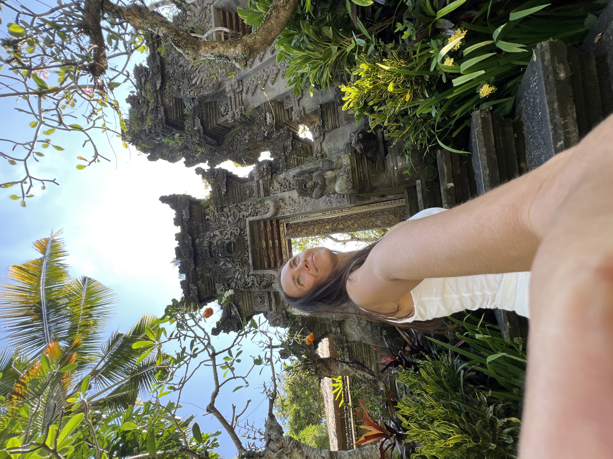 A medium shot depicts a woman in a white tank top, standing and taking a selfie in front of a traditional Balinese stone structure in Kedewatan, Indonesia. The structure is ornately carved and partially obscured by foliage. The sky is visible through openings in the structure and foliage. Tropical plants and a palm tree are visible on the left side of the image. The lighting is bright, with some shadows cast by the foliage.