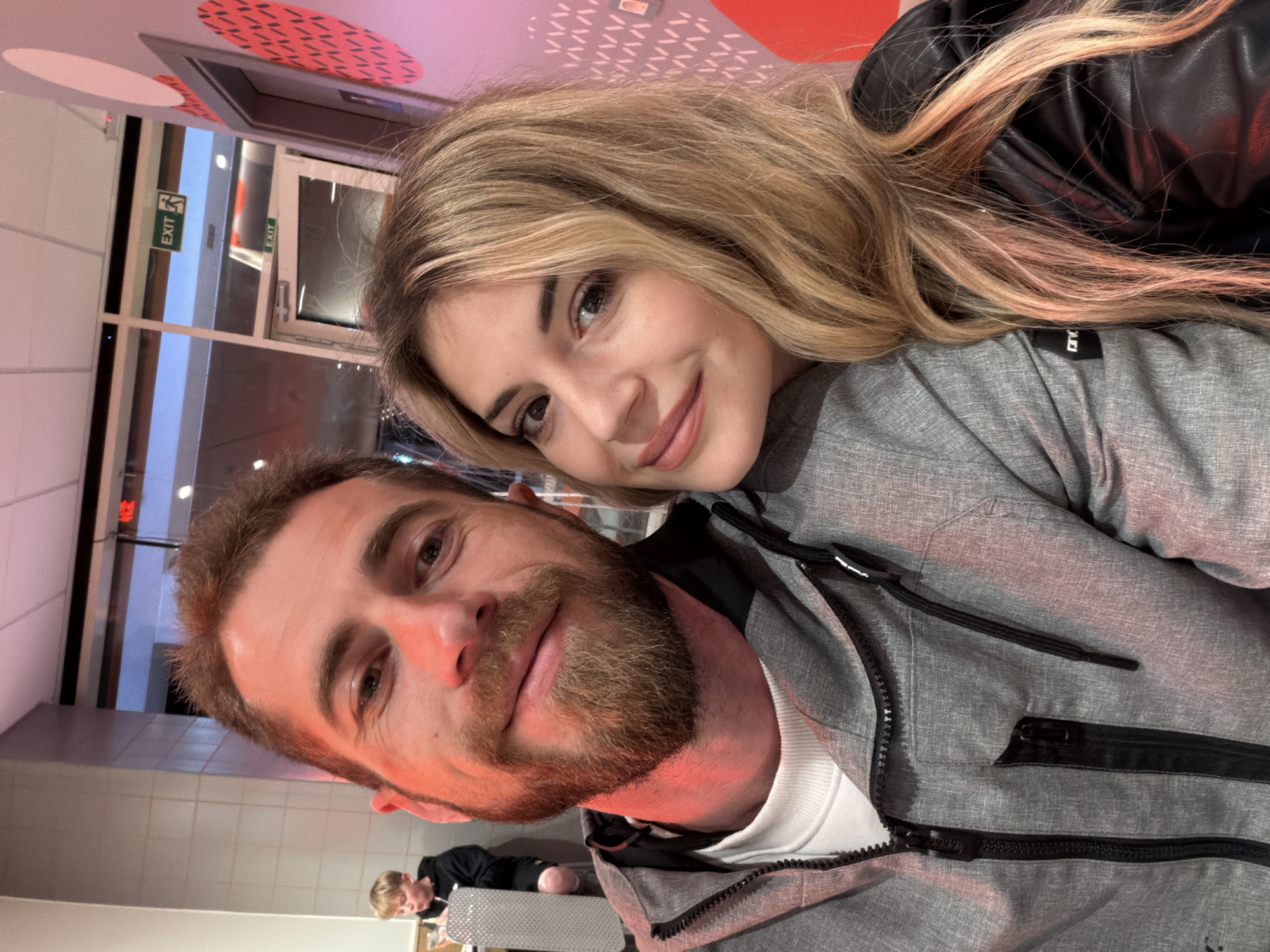 A tilted, close-up selfie captures a man and a woman in an indoor setting, likely a public space such as a restaurant or shopping center in Budva, Montenegro. The image is rotated approximately 90 degrees counter-clockwise.
The woman, with long blonde hair, is positioned in the upper part of the frame, leaning into the man. She wears a grey top and a black leather jacket. Her face is turned towards the camera, adorned with subtle makeup and a gentle smile, revealing dark eyes.
The man, in the lower part of the frame, has a beard and light brown hair. He is wearing a grey zip-up jacket over a white top. His face is also directed towards the camera, with a slight smile. A striking reddish artificial light illuminates parts of his face and neck, creating a distinct color cast.
In the background, visible through glass panels or windows, is an illuminated "EXIT" sign with a green pictogram. Another "EXIT" sign is faintly visible on a doorframe. The background also shows light-colored walls, possibly tiled, and a ceiling decorated with red and white geometric patterns. In the lower left corner, another person, appearing younger, is partially visible sitting at a table with a reflective surface. The overall atmosphere suggests an evening or night setting due to the artificial lighting. Both individuals appear content and happy, sharing a moment captured in the selfie.