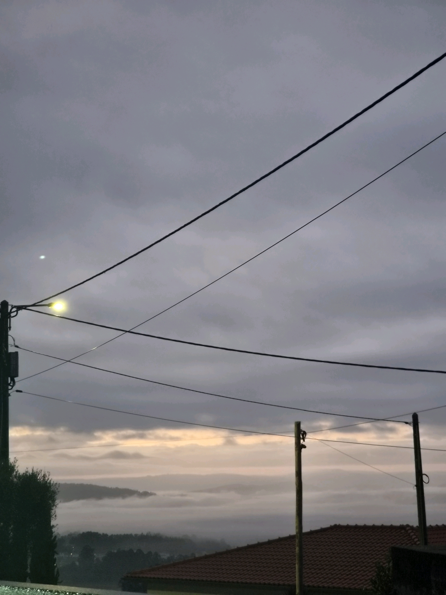 The image captures a misty landscape in Barcelos, Portugal, at dusk. Multiple dark electrical wires crisscross the frame, supported by a utility pole on the left with a lit lamp and a smaller, partially visible box. Two additional wooden utility poles are visible in the mid-ground, supporting more wires. In the background, a hazy, ethereal layer of mist blankets rolling hills, with hints of a soft orange glow from the setting sun peeking through the clouds at the horizon. The foreground shows the edge of a terracotta-tiled roof and the silhouette of a dark green tree on the left. The overall atmosphere is subdued and peaceful, illuminated by the dim, overcast light.