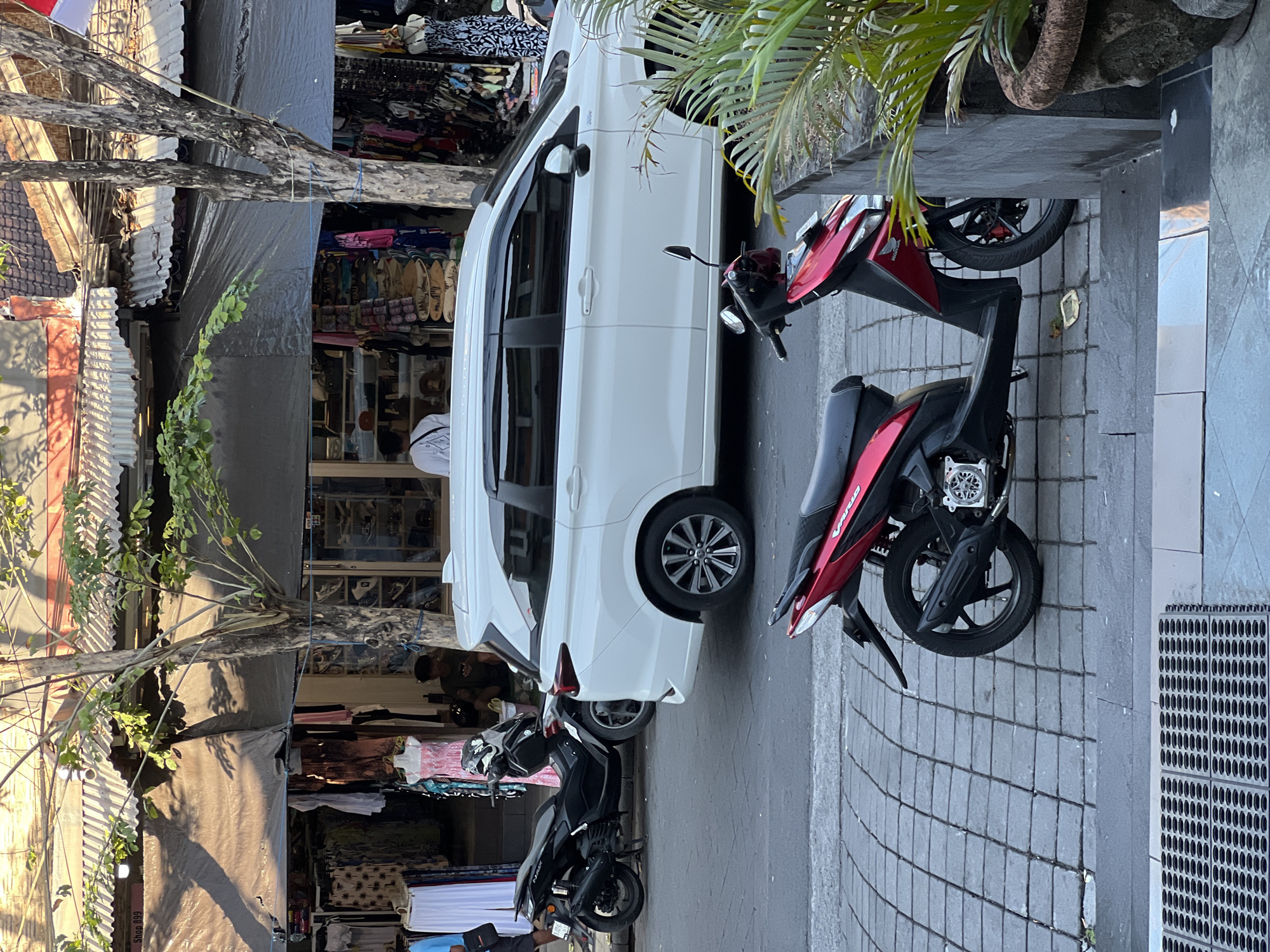 In Kuta, Indonesia, on a street lined with shops, a white minivan is parked on the road. In front of the minivan, a black scooter is parked on the sidewalk. Another red and black scooter is parked on the sidewalk in the foreground. The shops display clothing and souvenirs. The sun casts shadows on the street and sidewalk.