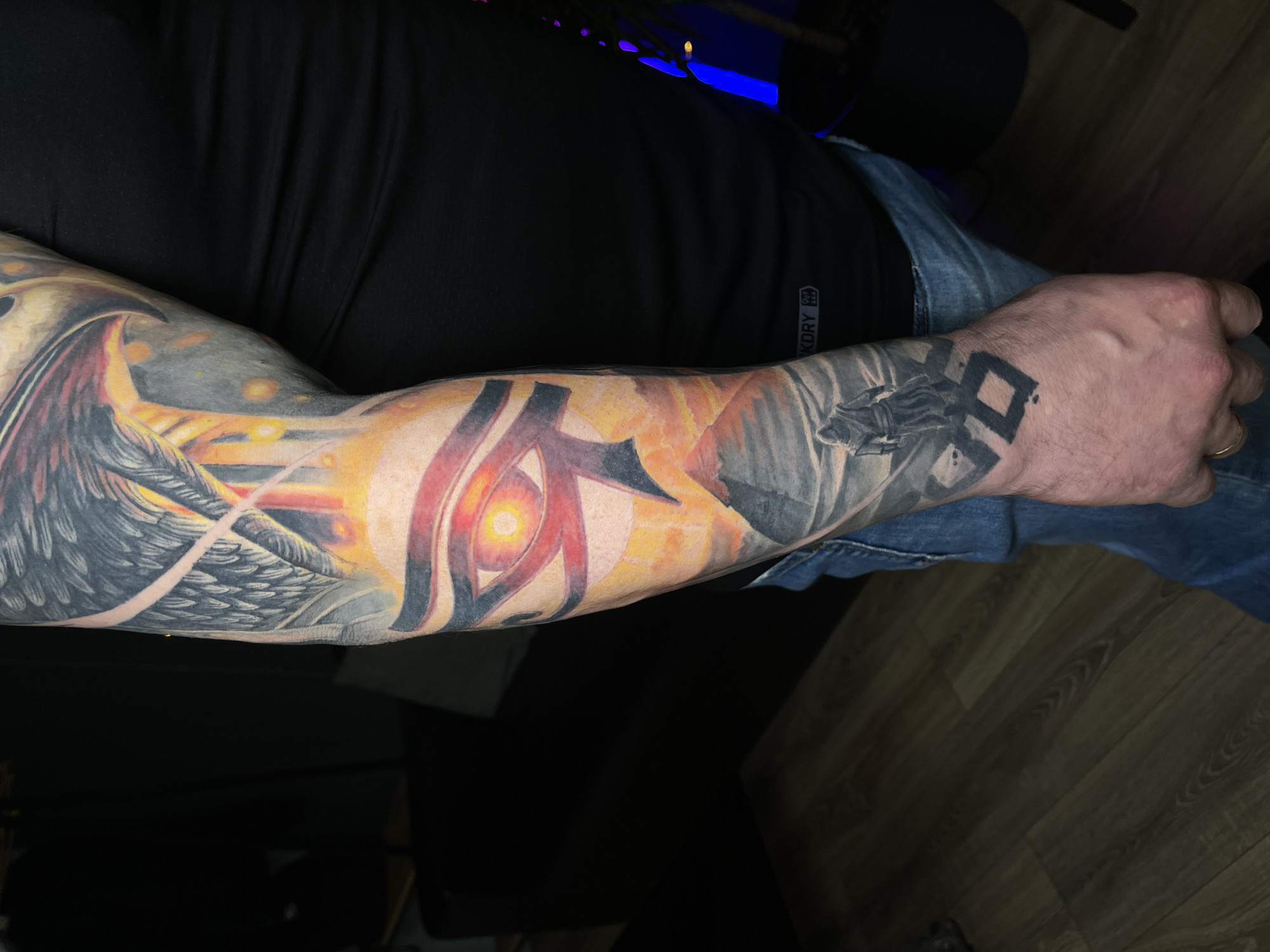 A person's arm is displayed, heavily tattooed. The tattoos include a large eye design, a winged figure, and geometric shapes on the hand. The person is wearing a black shirt and blue jeans. The setting appears to be indoors with a wooden floor. The location is not evident from the image.