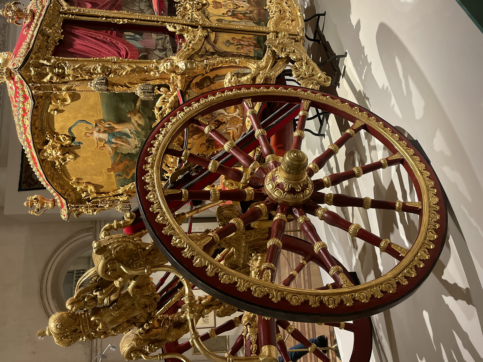 The image shows a highly ornate, gold-trimmed, red-wheeled carriage, likely a royal or ceremonial coach. The wheel is prominently displayed in the foreground, with detailed gold embellishments around its rim and center. The carriage's body is richly decorated with gold carvings and painted panels depicting scenes of figures. The environment appears to be a museum or historical exhibit. The location in the city of Saint Petersburg, Russia is implied due to the opulent style and historical context of the object.