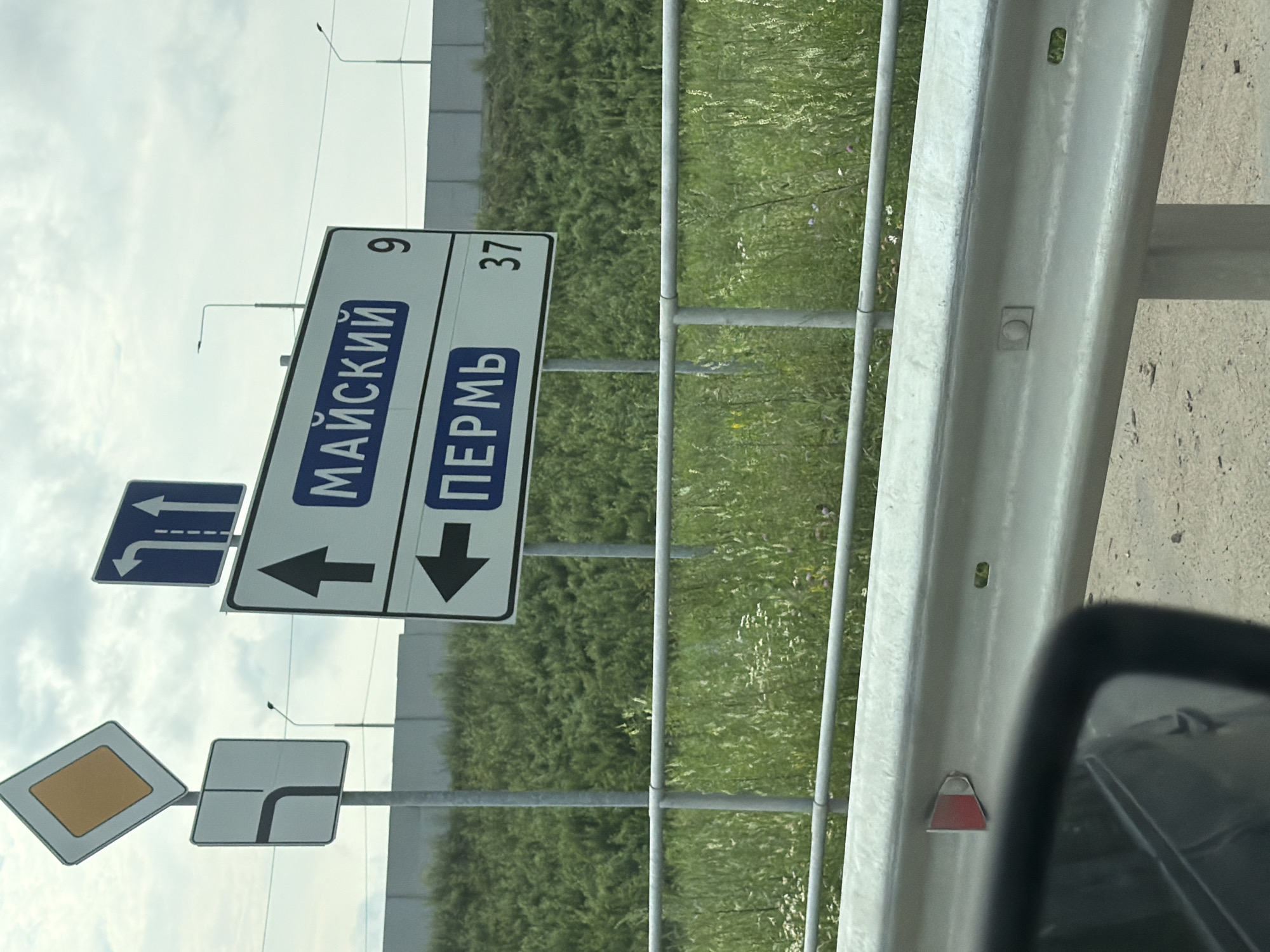 This image depicts a street scene with multiple road signs, viewed from a vehicle. The primary focus is on a set of directional signs. One large white sign with a black border indicates "МАЙСКИЙ" (Maysky) with the number "9" and "ПЕРМЬ" (Perm) with the number "37". Below these, black arrows point left and down, respectively. To the left of this sign, a blue rectangular sign shows two white arrows, one pointing left and the other indicating a right turn with a dashed line. Another diamond-shaped sign with a yellow interior is visible, along with a square sign showing a road turning right.
The setting appears to be a roadside with a metal guardrail in the foreground, separating the road from a grassy area with wildflowers and some trees. The sky is overcast with clouds, suggesting a daytime scene with possibly changeable weather. The overall impression is that of navigating a road in an urban or semi-urban environment, likely in or near Krasnokamsk, Russia, given the Russian text on the signs. The image is captured from inside a vehicle, as indicated by the partial view of a rearview mirror on the right side. There are no people visible in the image.