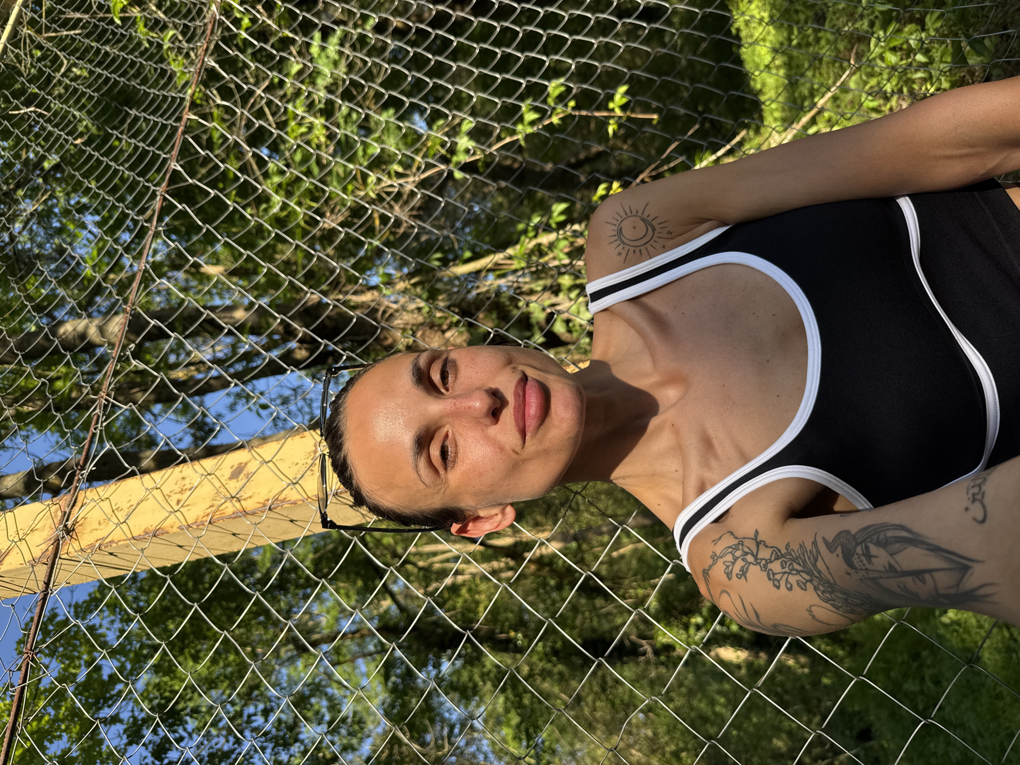 A woman with dark hair pulled back and glasses perched on her head stands in front of a chain-link fence. She wears a black tank top with white trim. Tattoos are visible on her upper arms; one features a sun and moon design, and another, a larger, more detailed image. The fence has a yellow metal post. Beyond the fence, trees and sky are visible. The picture was taken in Pleven, Bulgaria.