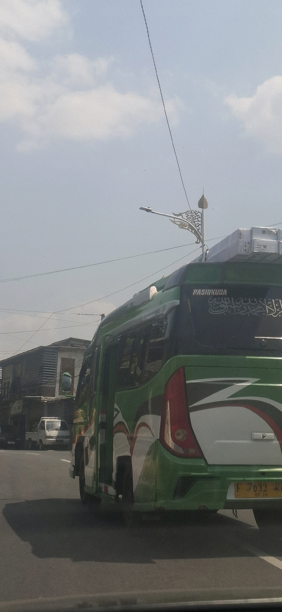 In Sukasari, Indonesia, a green and white bus is captured from a rear-side perspective, traveling on an asphalt road under a bright, partially clouded sky. The bus features a decorative Arabic script on its rear window, with the word "PASIRKUDA" above it. A white pickup truck is visible to the left of the bus, and behind it, a dark-colored truck is parked near a building. The environment suggests an urban or suburban setting with overhead electrical wires and a lamppost with an ornate design. The time of day appears to be afternoon, indicated by the sunlight and shadows cast on the road. The bus has a license plate reading "F 7611 WK 07.26".