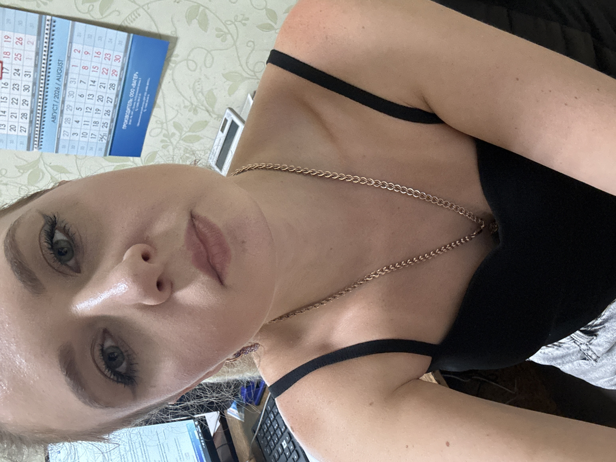 A close-up selfie of a young woman with blonde hair and green eyes. She is wearing a black tank top and a gold necklace. Her makeup includes eyeliner and mascara. In the background, a calendar shows August 2026. The overall setting appears to be an indoor office or room, likely in Makiivka, Ukraine, given the Cyrillic text on the calendar.