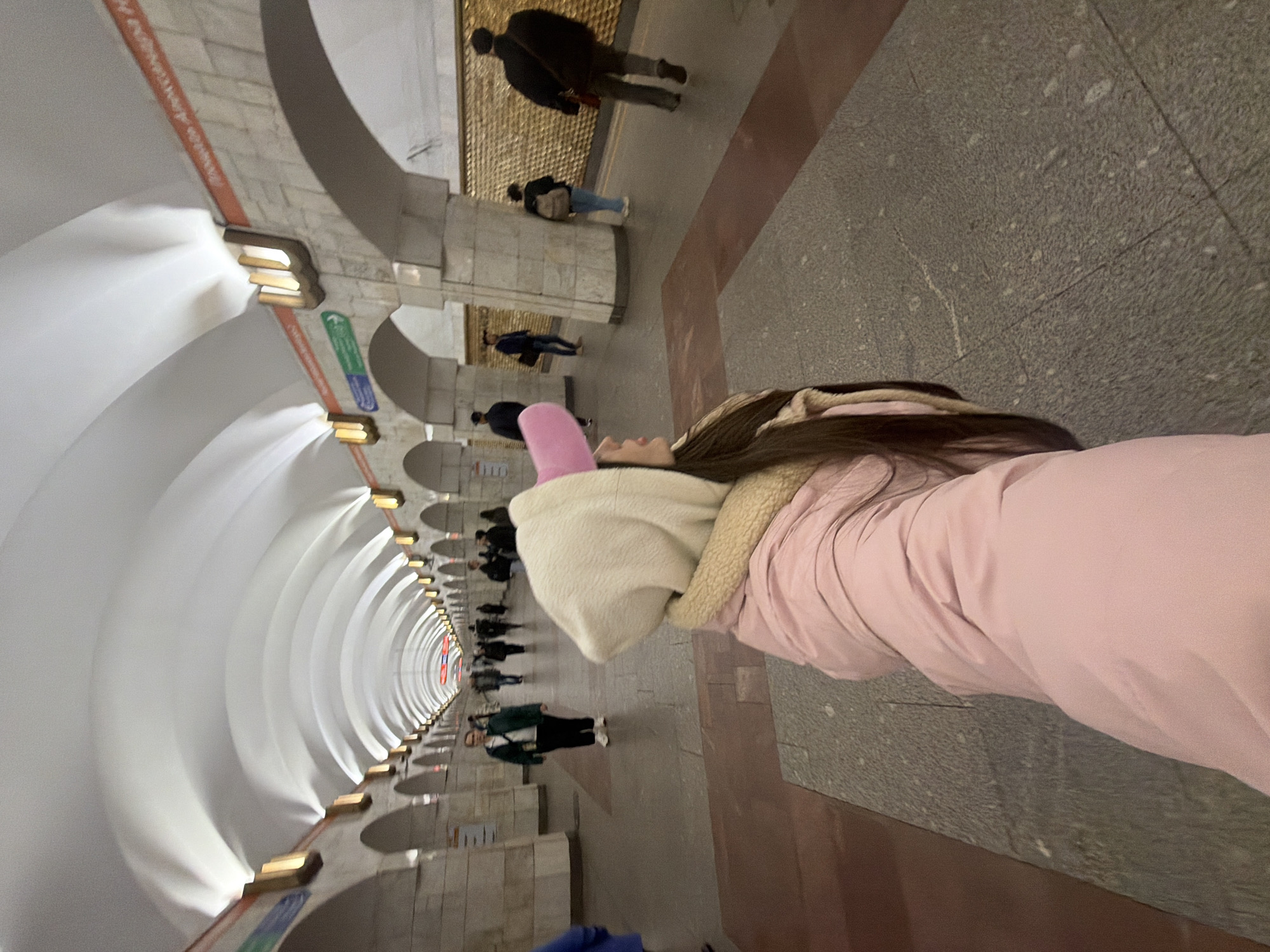 The image presents a dramatically rotated view of a grand underground subway station. The perspective is tilted sharply clockwise, as if looking up from a reclining position, creating a dynamic, almost disorienting, visual of the long tunnel.
In the right foreground, the upper body of a person is visible. They are wearing a light pink puffer jacket and a cream-colored hoodie adorned with distinctive pink "bunny ears." Their face is seen in profile, looking down the length of the subway tunnel.
The setting is characterized by a series of repeating white, vaulted arches that form a long, symmetrical tunnel. Warm-toned light fixtures are integrated into the side walls within these arches, casting a soft glow and highlighting the architectural curves. The platform below features pillars made of light-colored stone or concrete. The floor is covered with large, mottled grey tiles, interspersed with sections of reddish-brown. A reddish-orange stripe, bearing faint and illegible text, runs horizontally along the lower part of the wall.
Several other commuters are scattered along the platform at various distances, some standing, others seemingly in motion. They appear mostly blurred, emphasizing the depth of the tunnel. A small sign on one of the pillars displays a green arrow pointing right, with a blue rectangular shape below it. At the far end of the tunnel, a distant red and white circular logo, possibly a metro emblem or line indicator, is faintly visible.
The primary activity depicted is the person in the foreground seemingly taking a photo or selfie, capturing the impressive architecture of the subway station. The overall atmosphere is one of well-lit, functional grandeur typical of older metro systems.
