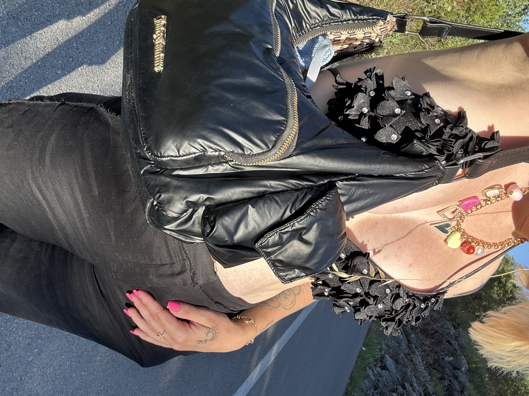 A person stands on what appears to be an asphalt surface, likely a road or sidewalk. The person is wearing black jeans, a black top with flower-like embellishments near the collar, and a black puffer-style crossbody bag. The bag features a gold-toned logo. The person's hand is visible, with pink nail polish. A gold ring and bracelet are also visible on the hand and wrist. The person has blonde hair and is wearing a necklace with colorful pendants. A tattoo is visible on the forearm. The location appears to be outdoors, with a rocky area and vegetation visible in the background. A road marking is also visible on the asphalt. The photo is taken from a top-down perspective and is rotated slightly. The specific location in Dubraljevina, Montenegro, is not discernible from the image.