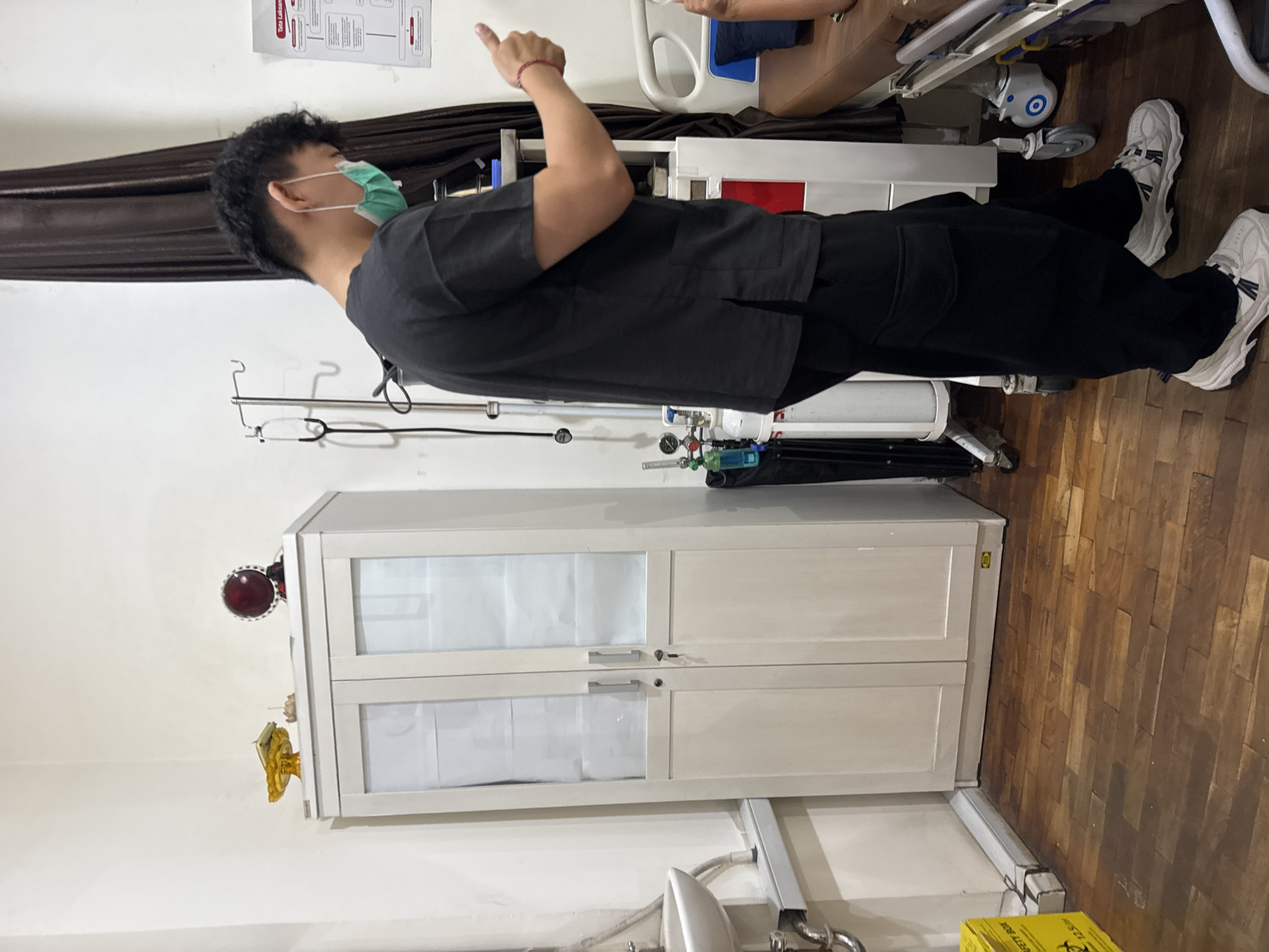 A person wearing a medical mask, dark scrubs, and white sneakers stands in what appears to be a medical facility or simulation lab in Kuta Selatan, Indonesia. They are giving a thumbs-up gesture. An IV stand, medical equipment (possibly oxygen tanks), and a white cabinet with translucent doors are visible. A medical training mannequin is partially seen in the background. The room has white walls and a wooden floor.