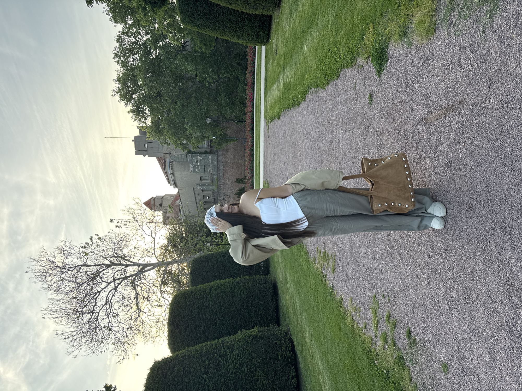 A woman stands on a gravel path, slightly turned towards the left, in what appears to be a park setting. She is wearing a white top, green pants, a light beige jacket draped over her shoulder, and white shoes. She also has a white head covering. She is holding a tan-colored bag with studded details. The backdrop includes manicured hedges, green grass, and a medieval-style stone building or castle under a bright, overcast sky. A bare tree is partially visible on the left.
This is NOT in Belgrade, Serbia. The castle is Malmöhus Castle in Malmö, Sweden.