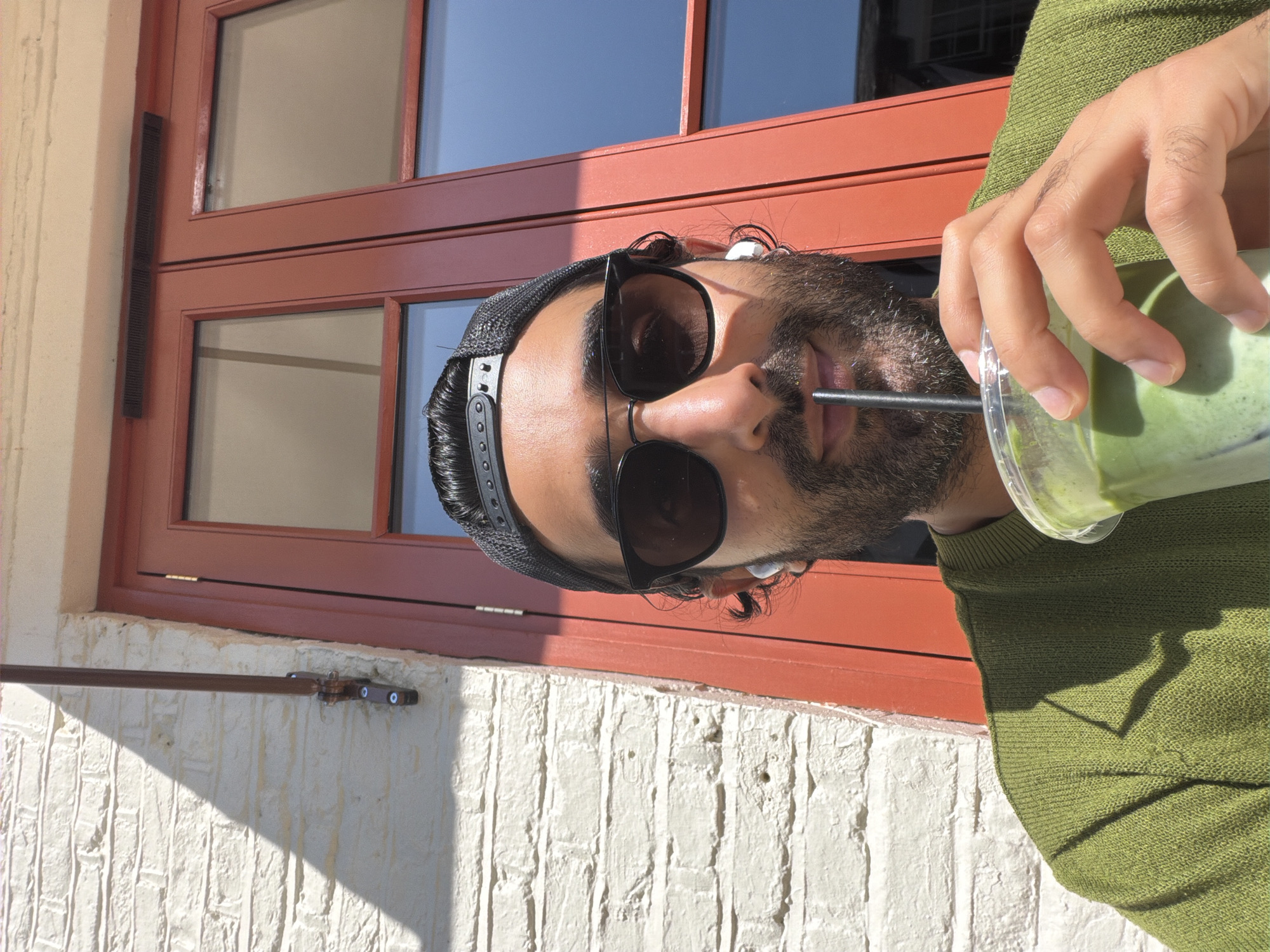 A man wearing a green sweater and a black baseball cap is drinking a green beverage from a clear plastic cup with a black straw. He has on black sunglasses and white earbuds. He is outdoors in front of a red door and a white brick wall. A shadow is cast on the wall from above. The scene appears to be in London, United Kingdom.