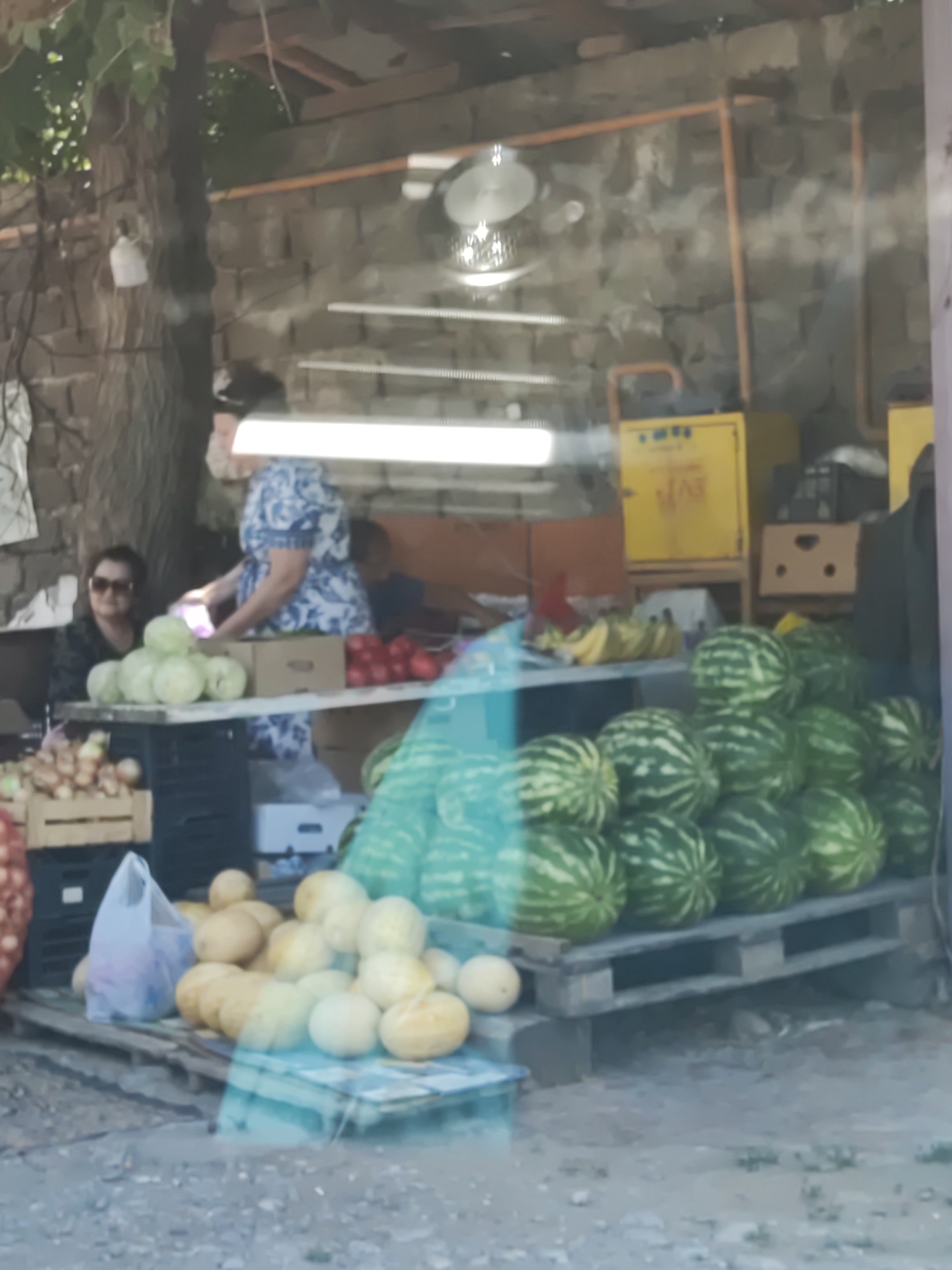The image depicts an outdoor market stall likely located in Shymkent, Kazakhstan, with a focus on fresh produce. A woman in a blue and white patterned dress is standing behind a table, seemingly attending to customers or organizing goods. To her left, another woman wearing sunglasses is seated. The stall displays a variety of fruits and vegetables, including a large pile of watermelons stacked on wooden pallets, several cabbages, onions in crates, and a bunch of bananas. Behind the stall, a rustic stone wall forms the backdrop, with some shelving and yellow storage units visible. The overall scene suggests a typical day at a local marketplace.