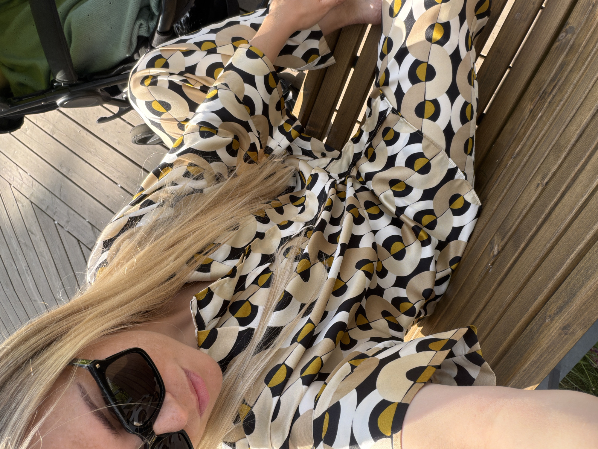 A young female individual is seated on a slatted wooden surface in Votkinsk, Russia. She has long, straight blonde hair and fair skin with visible freckles on her nose and cheeks. She wears oversized black "Polaroid" brand sunglasses. Her attire consists of a matching two-piece set, a collared long-sleeved shirt and trousers, featuring an abstract geometric pattern of interlocking and overlapping circles and semi-circles in shades of beige, cream, black, and mustard yellow. The fabric exhibits a sheen suggestive of silk or satin. Her right arm is bent, and her bare feet are visible, resting on the wooden planks. To her right, a black-framed baby stroller with a green blanket draped inside is partially visible. The scene is illuminated by strong, direct sunlight, casting distinct shadows on the wooden surface, which appears to be a deck or bench.