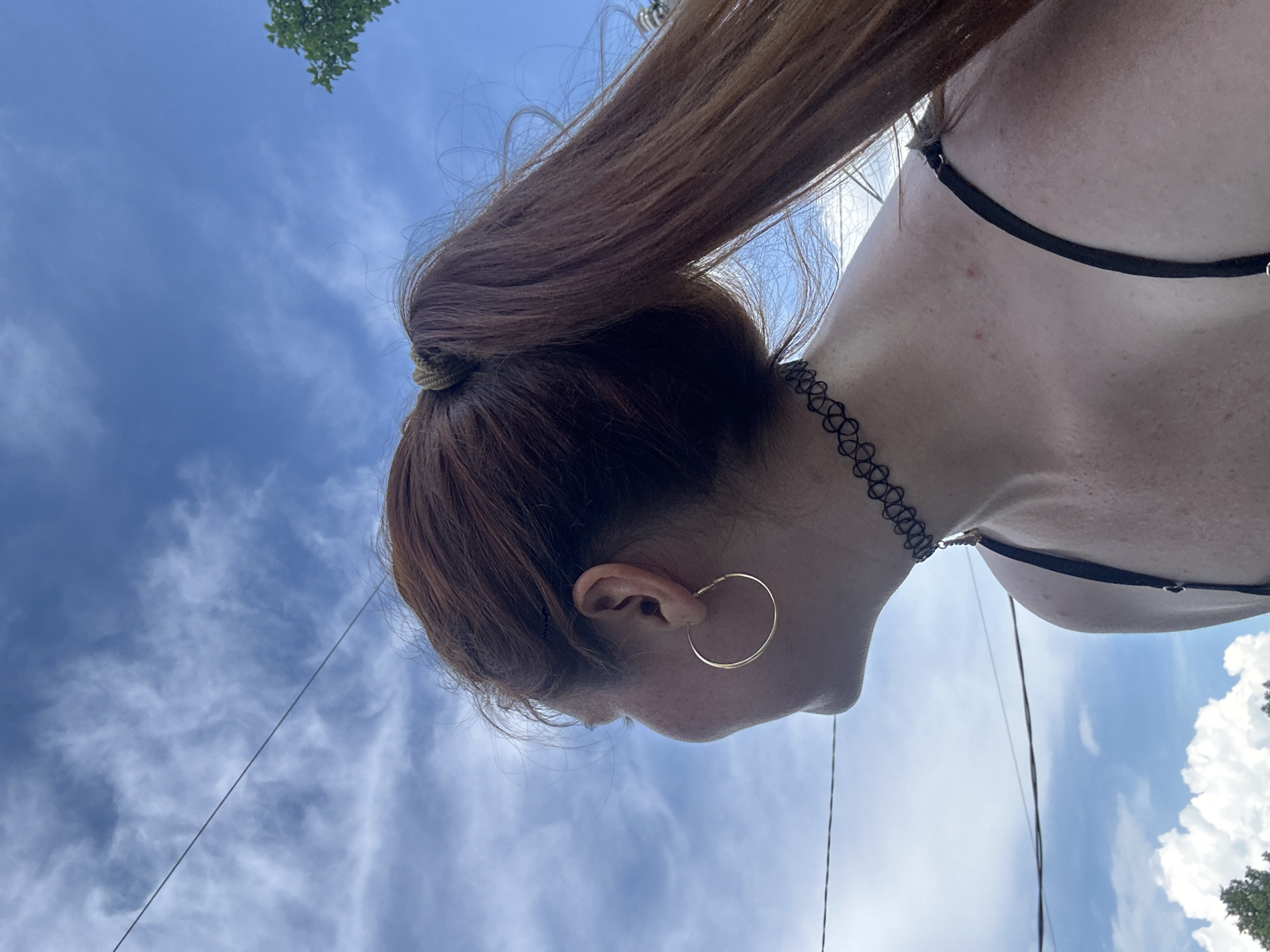 A medium shot image shows the back of a person's head and shoulders against a blue sky with white clouds. The person has long, auburn hair pulled into a ponytail. They are wearing a black choker necklace and a black spaghetti strap top. A thin gold hoop earring is visible in their left ear. The image is taken outdoors, and there are power lines visible in the upper portion of the frame, along with a distant green tree branch. There is no indication of the location being Nizhny Novgorod, Russia.
