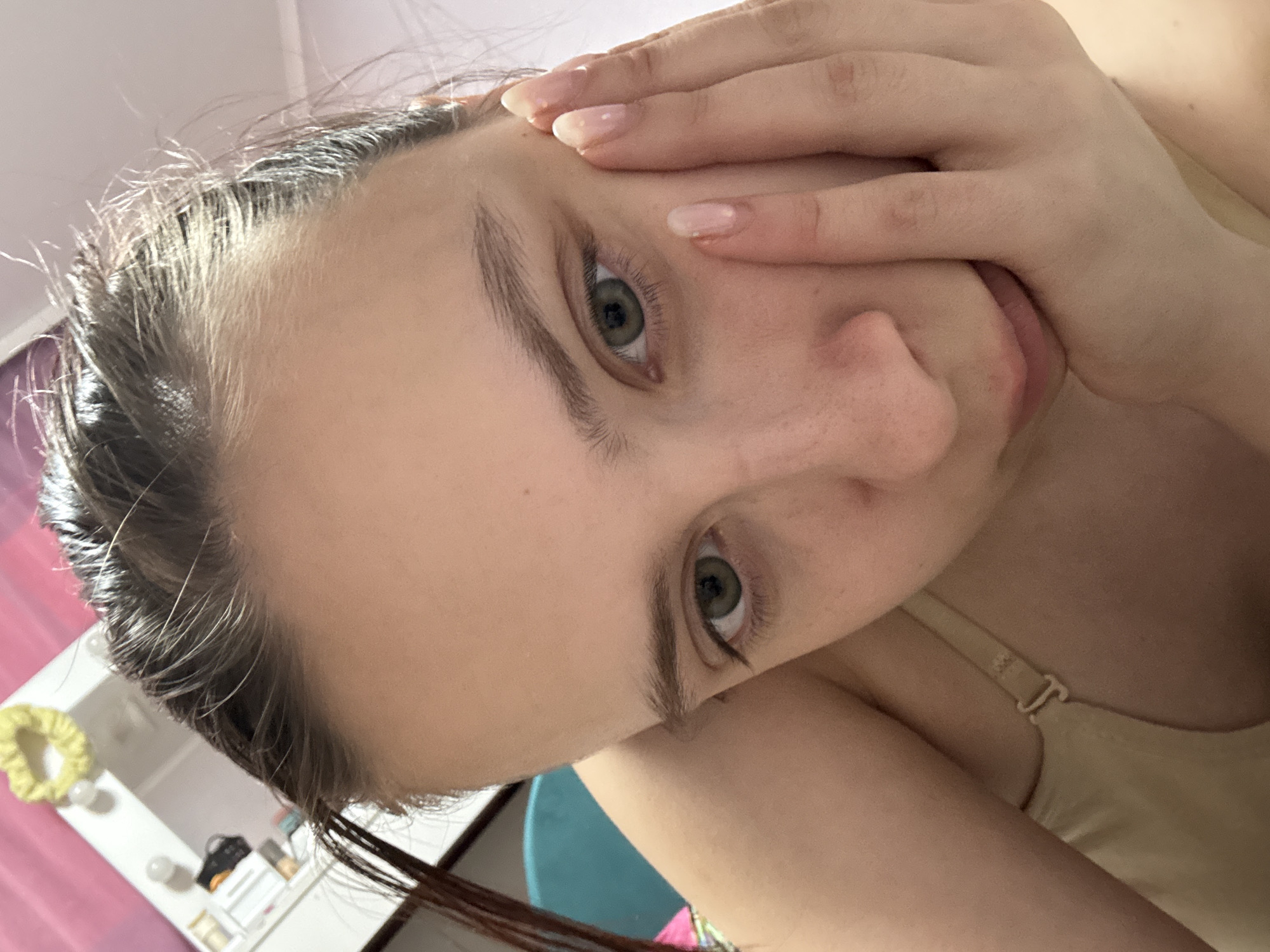 A close-up photograph shows a person's face partially obscured by their hand. The individual has fair skin, light eyebrows, and green eyes. The hair appears wet or damp. The hand is positioned near the face, with fingers partially covering the nose and mouth. The person is wearing a beige bra. The background includes a blurred image of a shelf and a teal object. The location, Прикамский, Russia is not discernible from this image.