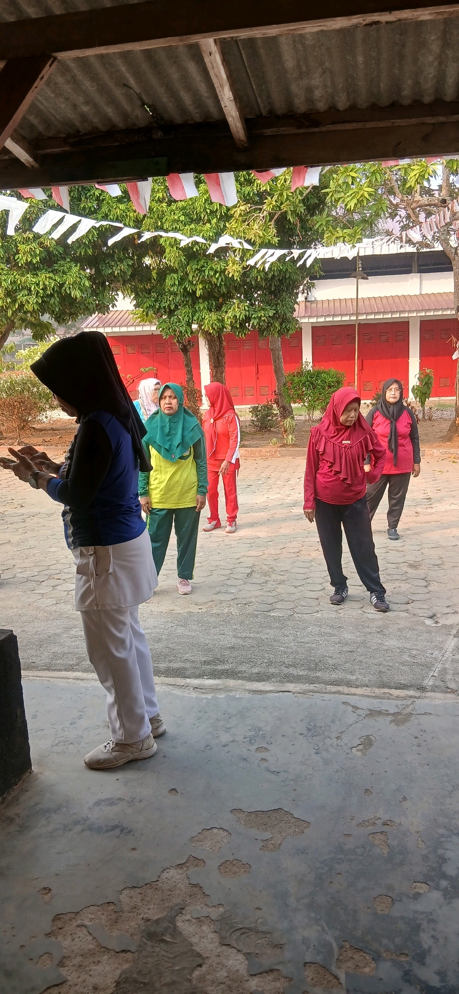 In the city of Metro, Indonesia, a group of women is engaged in physical activity outdoors, possibly a form of exercise or a community gathering. The scene is set under a weathered, corrugated metal roof supported by wooden beams. Red and white Indonesian flags, strung as bunting, hang decoratively from the roof structure and extend across the background, suggesting a festive or patriotic occasion.
The women are dressed in modest athletic attire, predominantly hijabs and tracksuits in various colors including green, yellow, red, and black. One woman in the foreground, on the left, wears a black hijab and a blue long-sleeved shirt with white accents on her pants. She appears to be looking down at something in her hands. Behind her, a woman in a green hijab and a yellow and green tracksuit stands with a focused expression. To her right, another woman in a red hijab and red tracksuit stands with her hands on her hips. Further to the right, a woman in a maroon hijab and tracksuit is also present. The ground is paved with interlocking hexagonal stones, showing signs of wear and tear in some areas. In the background, a building with closed red doors is visible, flanked by trees with green foliage. The lighting suggests it is daytime, likely morning or late afternoon, given the angle of the shadows. The overall atmosphere appears to be one of organized recreation and community participation.
