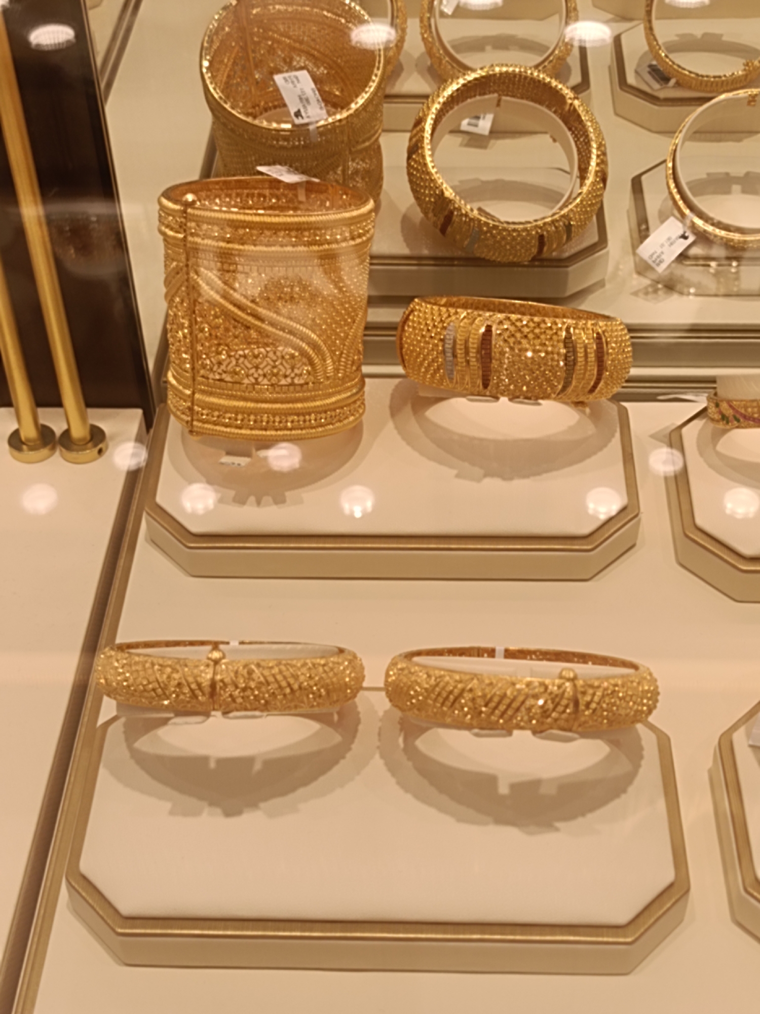 The image depicts a display of gold jewelry, specifically bangles and bracelets, inside a store. The jewelry is arranged on white, octagonal stands with gold trim. The lighting is bright, creating reflections on the gold. Some of the items have small white tags attached. In the background, there are more gold jewelry items and display cases, suggesting a retail environment. The presence of gold jewelry in a display case with price tags implies a commercial setting. Given the prevalence of gold souks and high-end jewelry stores in Dubai, this image could be from a jewelry shop in locations such as the Gold Souk in Deira or a luxury mall like The Dubai Mall or Mall of the Emirates, all situated within Dubai, United Arab Emirates. The image is a still shot, with no people or actions depicted.