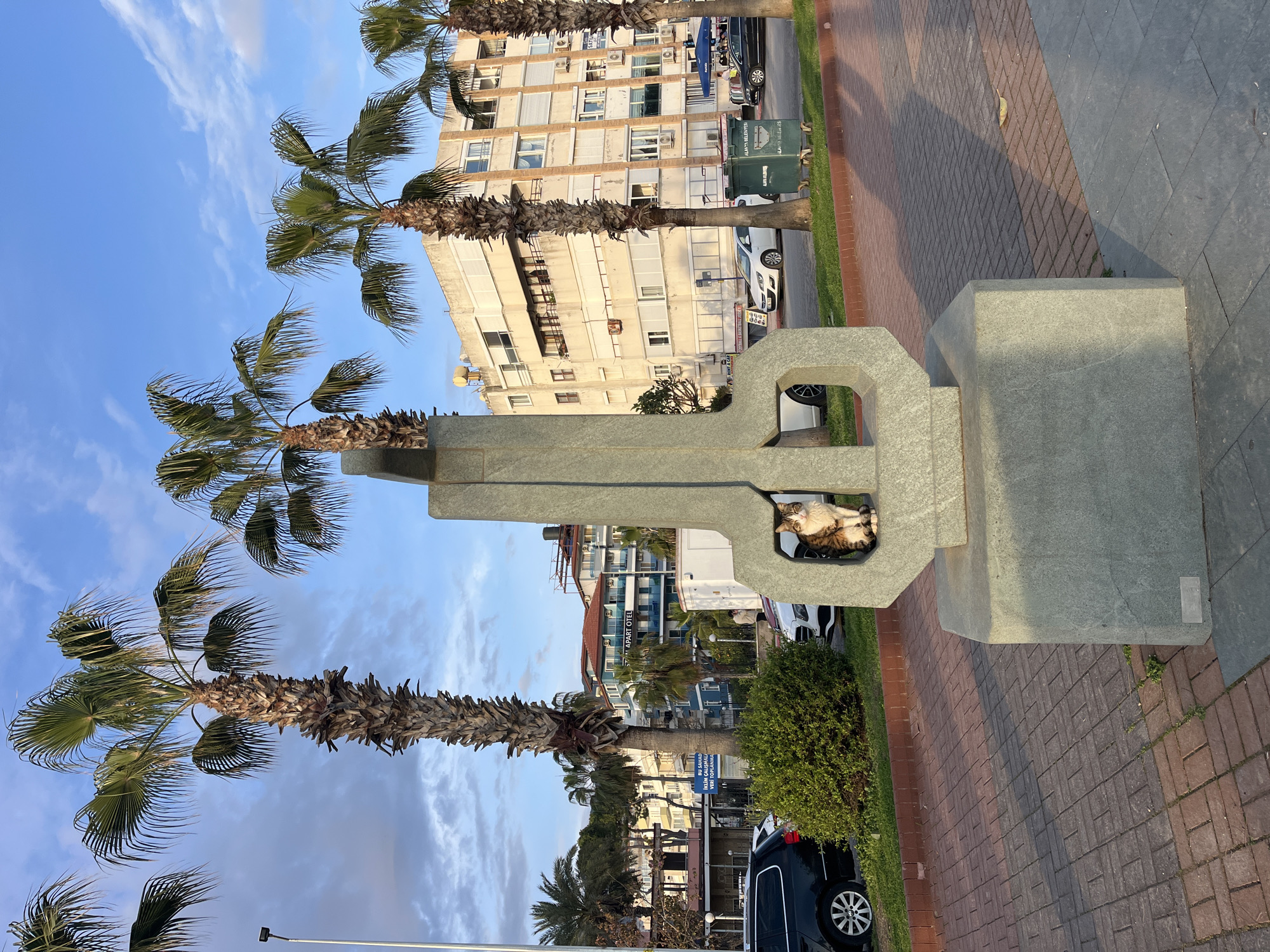 The image depicts a public space in Alanya, Turkey, recognizable by the city's characteristic palm trees and building architecture. A gray, concrete sculpture, possibly a monument or public art installation, stands prominently in the foreground. Within a square opening near the top of the sculpture, a cat is resting. The background includes an apartment building, several cars parked alongside the street, and a glimpse of other buildings and businesses. The foreground features a paved walkway, likely made of brick, and some greenery, suggesting a park-like or promenade setting. The sky is blue with light cloud cover.