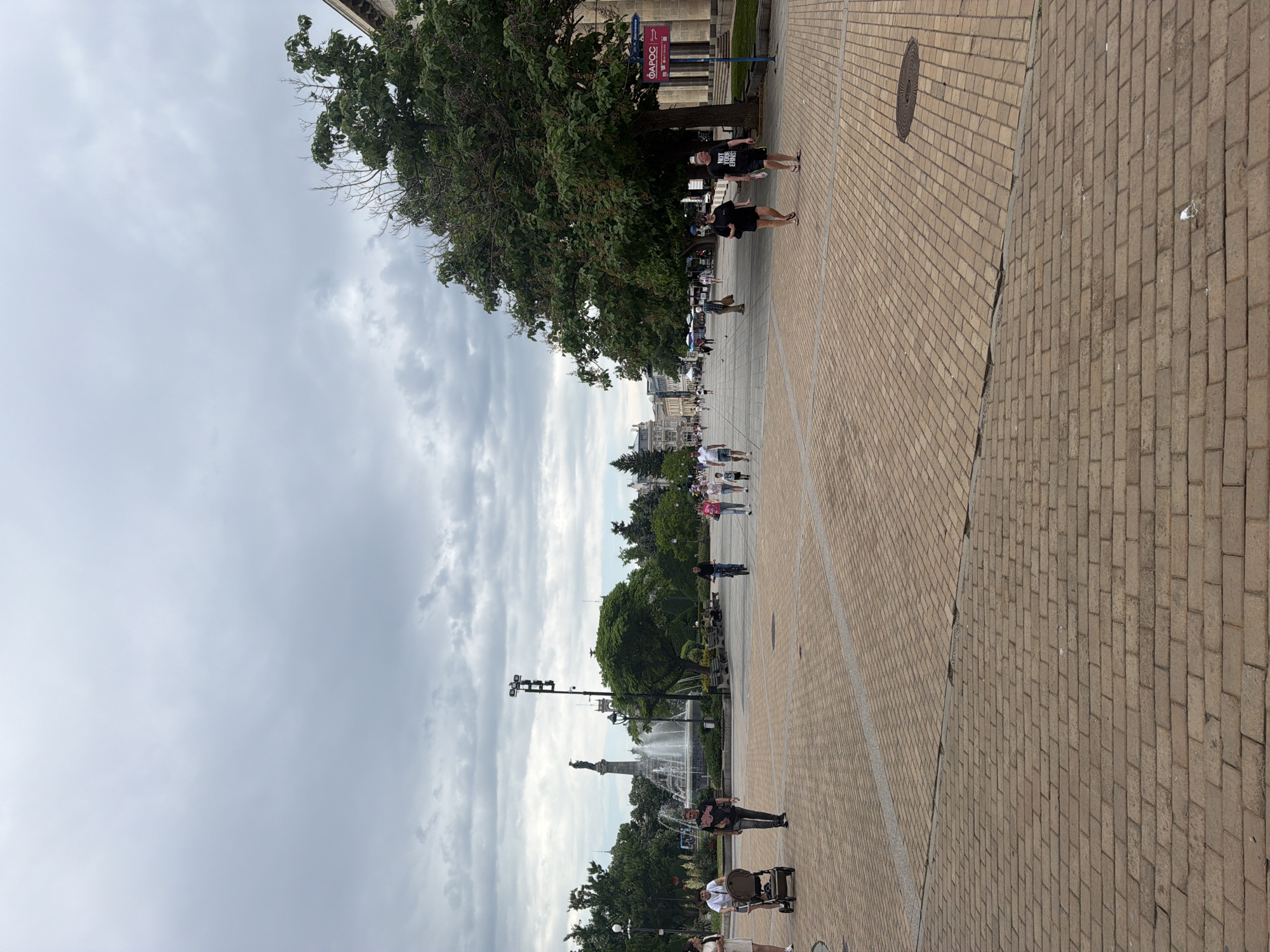 The image shows a wide, brick-paved public square in Ruse, Bulgaria, under a cloudy sky. People are walking along the square, and there are trees and bushes visible. A statue and a fountain can be seen in the middle distance. A sign written in Bulgarian is on the upper right. A manhole cover is on the ground in the upper right.