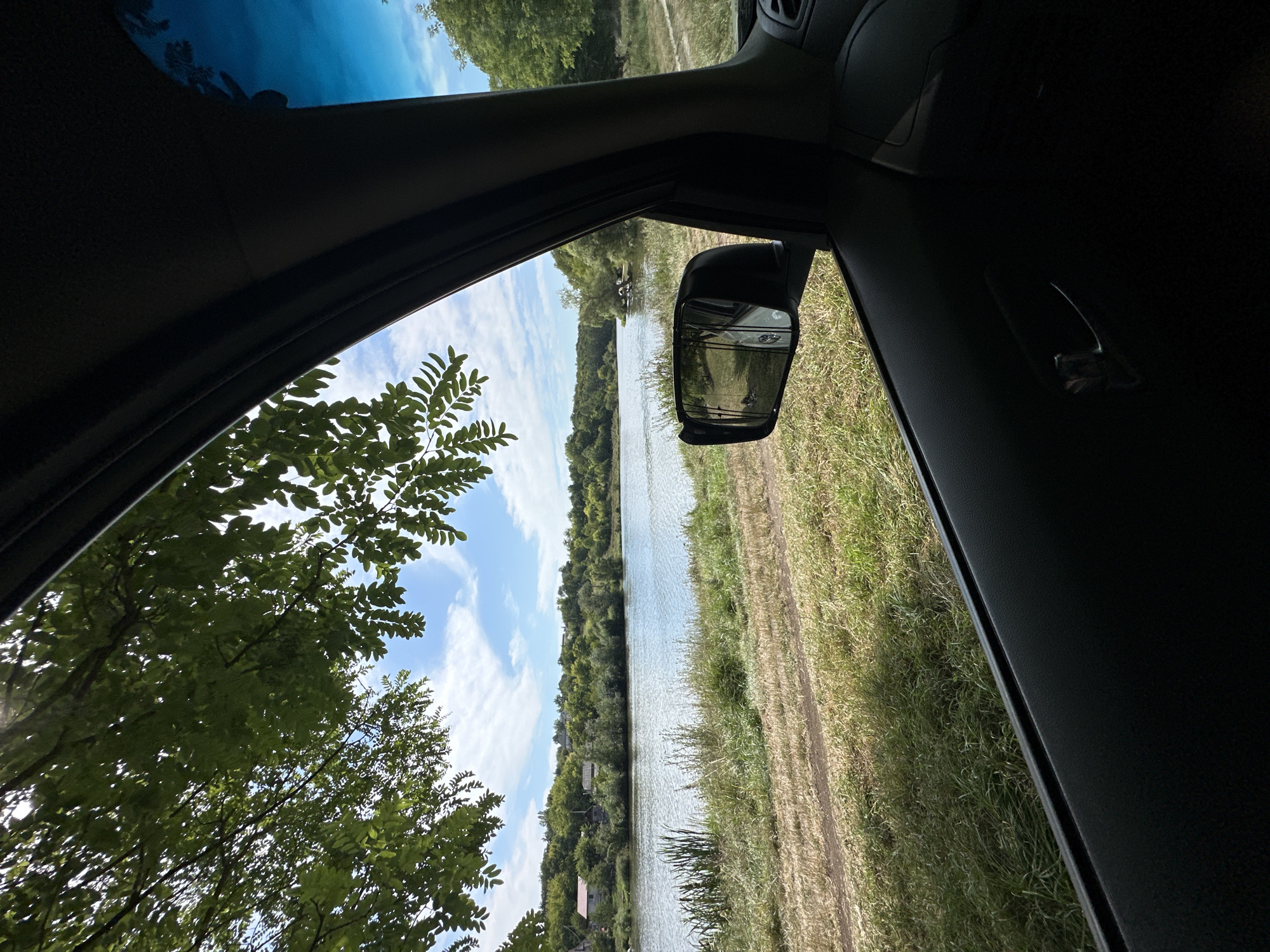 A view from inside a car looking out the passenger window towards a body of water bordered by a grassy bank and trees. The sky is blue with scattered clouds. The car's interior, including the door panel and side mirror, is visible in the foreground and to the right. The side mirror reflects a portion of the car's interior and the scene outside. The location is Makiv, Ukraine.