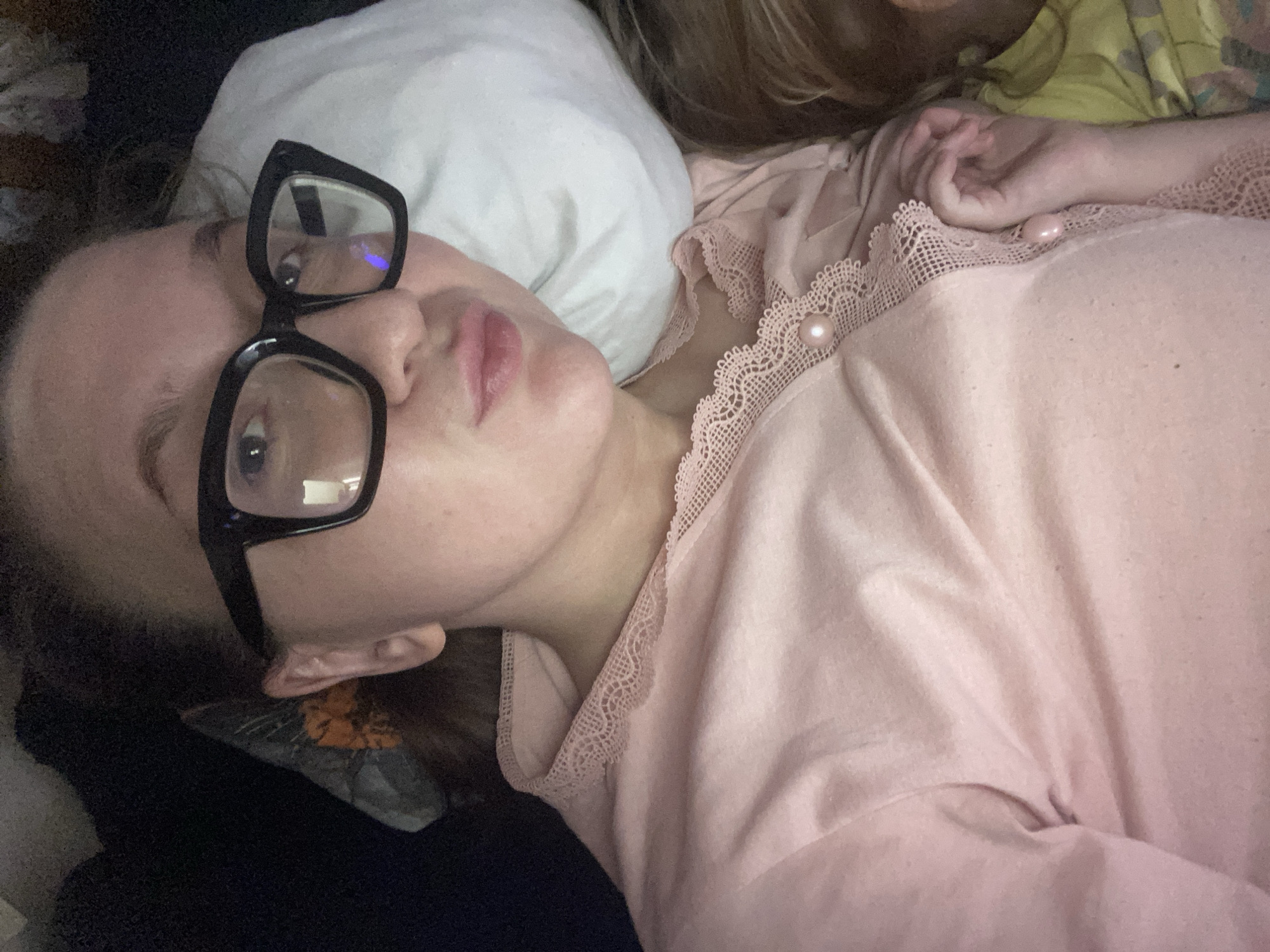 The scene depicts an indoor setting in Chaikovsky, Russia, featuring an adult female and a partially visible child. The adult female is positioned in a selfie-style view, lying supine. She wears black-framed eyeglasses and a light pink long-sleeved top with a lace-trimmed neckline and a front placket secured by at least one visible pearl-like button. Her lips are slightly pursed. Her dark hair is partially visible. A white pillow supports her head. To the lower right, the top of a child's head, with lighter hair, and a portion of a yellow patterned garment are visible, positioned adjacent to the adult's torso. The child's right hand is also visible. Dark, indistinct bedding or upholstery is present in the upper left background. The overall lighting is subdued.