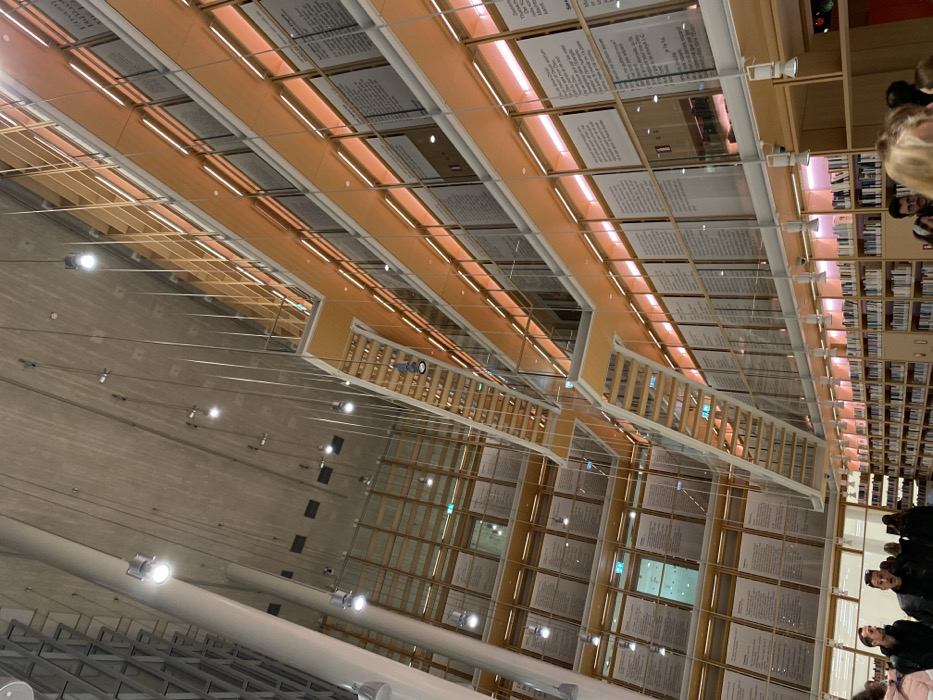 The image captures the interior of a large, modern architectural space, likely a library or cultural center, seen from a low-angle perspective looking upwards. The dominant features are the high ceilings and multi-level structures.
The ceiling, in the upper portion of the image, is a light, neutral color, possibly concrete, and features numerous bright spotlights. Spanning across the upper levels are long, rectangular panels illuminated from behind by warm strip lights. These panels contain dense blocks of text, resembling large pages from a book or document. While the specific content is illegible, the presence of extensive written passages is clear.
Beneath these text panels, light-colored wooden structures, possibly staircases, walkways, or shelving units, feature slatted railings. These structures create a sense of depth and multiple levels within the building. On the far right, extensive bookshelves filled with books are visible, confirming the setting as a library. Several people are visible in the lower right and far right of the frame, their heads and upper bodies suggesting they are standing and observing the space. Their expressions are neutral or not clearly discernible.
The overall scene is one of a spacious and contemporary public building. The lighting is artificial, with numerous spotlights and warm glowing strips, suggesting an interior setting where natural light may be supplemented or it is evening. The modern design, with clean lines, wood accents, and large text displays, is characteristic of a contemporary cultural institution. Given the context of Kallithea, Greece, this is likely the Stavros Niarchos Foundation Cultural Center, which houses the National Library of Greece.