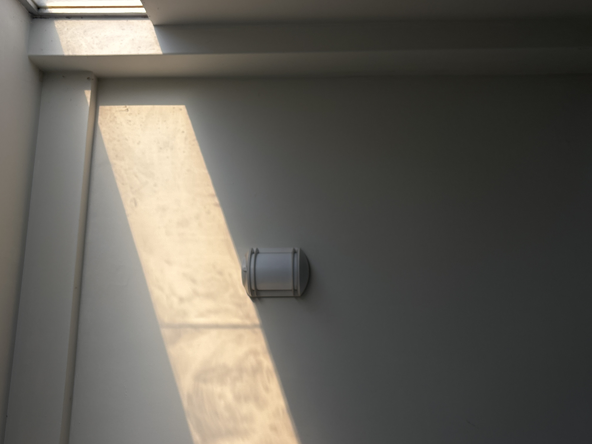 A harshly illuminated, white wall in Puebla, Mexico, is dominated by a strip of bright sunlight and a centrally located, cylindrical outdoor light fixture. The light is gray-white with circular ends and raised rings along its body. The wall, likely part of a building's exterior, is textured with a slight, grainy appearance. A vertical architectural element, possibly a column or wall extension, casts a shadow on the left side of the frame. A narrow strip of skylight appears at the top of the image.