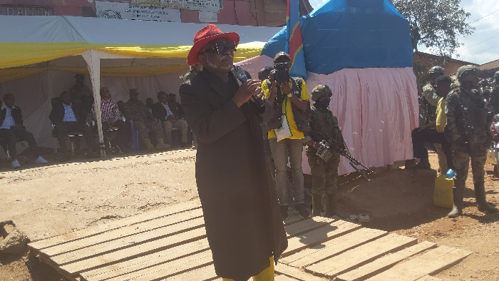 A man wearing a black jacket, a red hat, and sunglasses stands in front of a crowd and speaks into a microphone. There are several people sitting in chairs in the background under a white tent. A man with a camera is standing behind the man speaking and a group of military personnel are in the background on the right side of the frame. The event is taking place in Kirumba, Democratic Republic of Congo, at the coordinates -0.56 latitude and 29.179 longitude. A large blue and white tent is visible in the background with a pink drape. There is a wooden platform underneath the man speaking. The ground is dusty and the sky is clear. The man speaking appears to be the focal point of the scene.