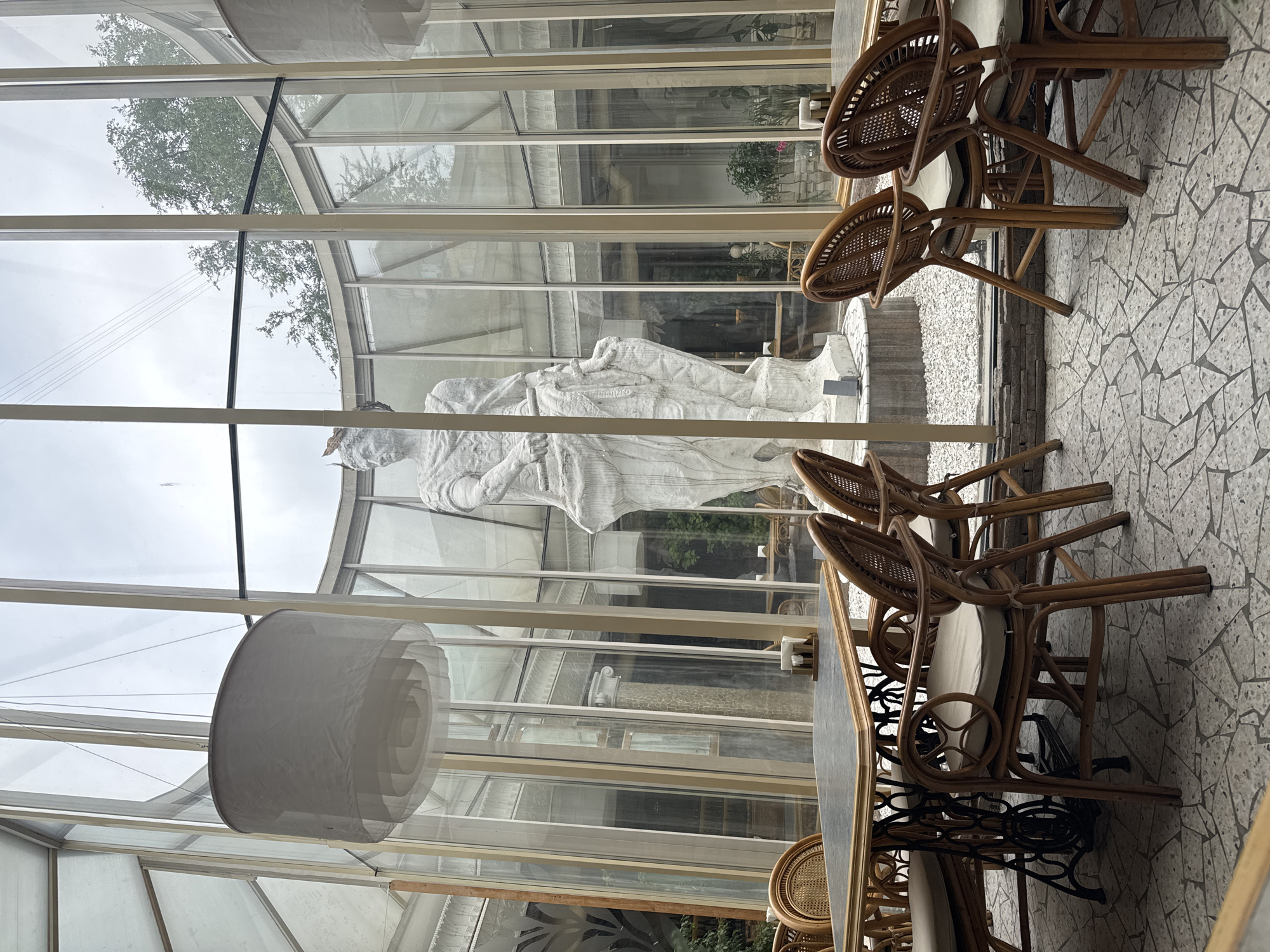 A large, white, neoclassical-style statue of a robed figure stands in the center of a sunroom, which features a curved glass exterior and a mosaic tiled floor. The statue is positioned on a white base. Several tables and chairs are arranged throughout the sunroom. In the foreground, a rectangular table with a dark top and an ornate, black metal base is surrounded by woven chairs with light-colored cushions. Another table and chairs are visible in the background, along with other architectural elements and greenery seen through the glass. A large, white, cylindrical lampshade hangs from the ceiling. The sky is overcast.