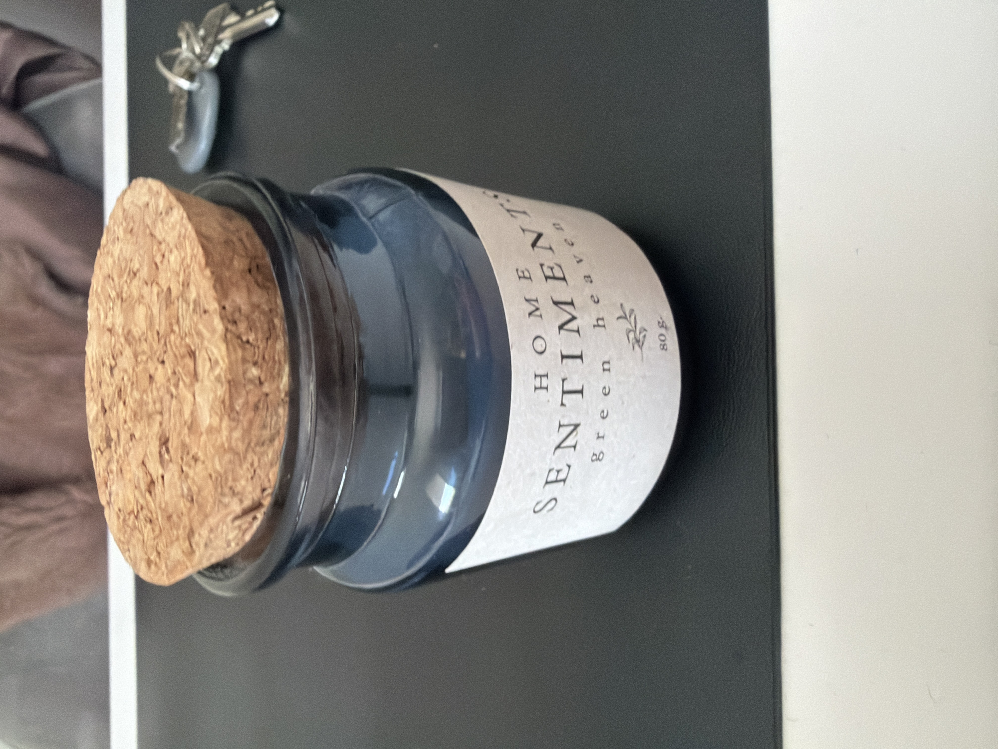 A blue glass jar with a cork stopper lies on a black surface next to a white surface. The jar has a label that reads "HOME SENTIMENTS green heaven 80g". A set of keys is visible in the upper left corner of the image. The scene provides insufficient information to determine if it is in Plovdiv, Bulgaria.