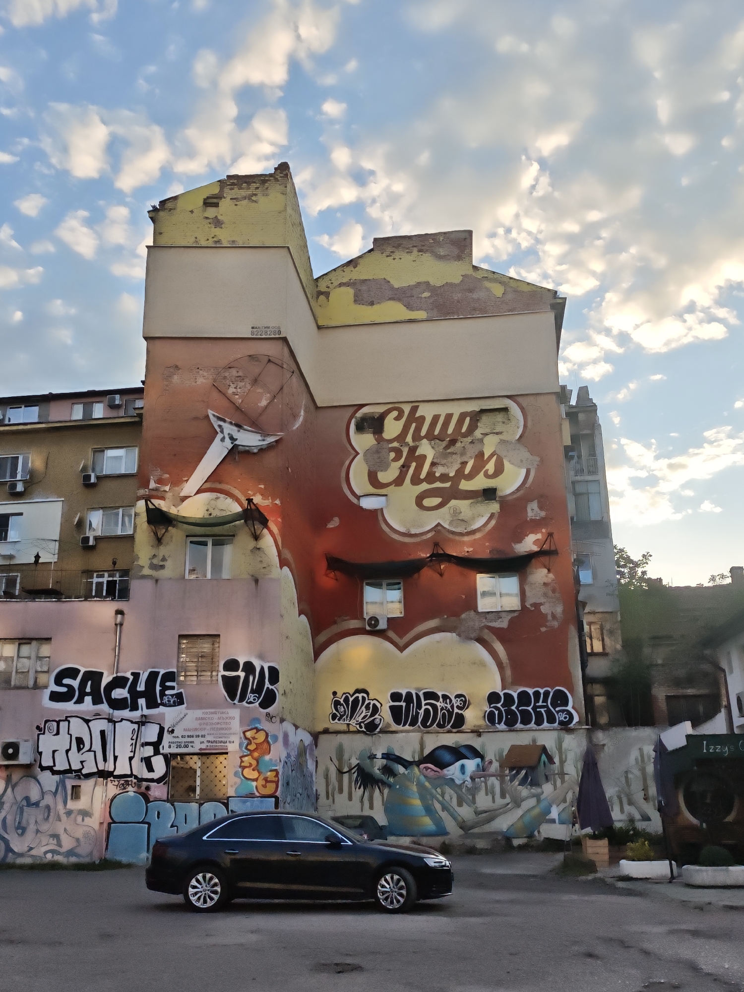 The image shows the exterior of a building in Sofia, Bulgaria. The building features graffiti, including faded Chupa Chups branding and a business sign. The graffiti style appears to be modern. A black car is parked in front of the building. The sky is visible above the building, with scattered clouds. The overall environment appears urban and possibly slightly rundown due to the graffiti and building's condition. Another apartment building is visible in the background behind the foreground building.