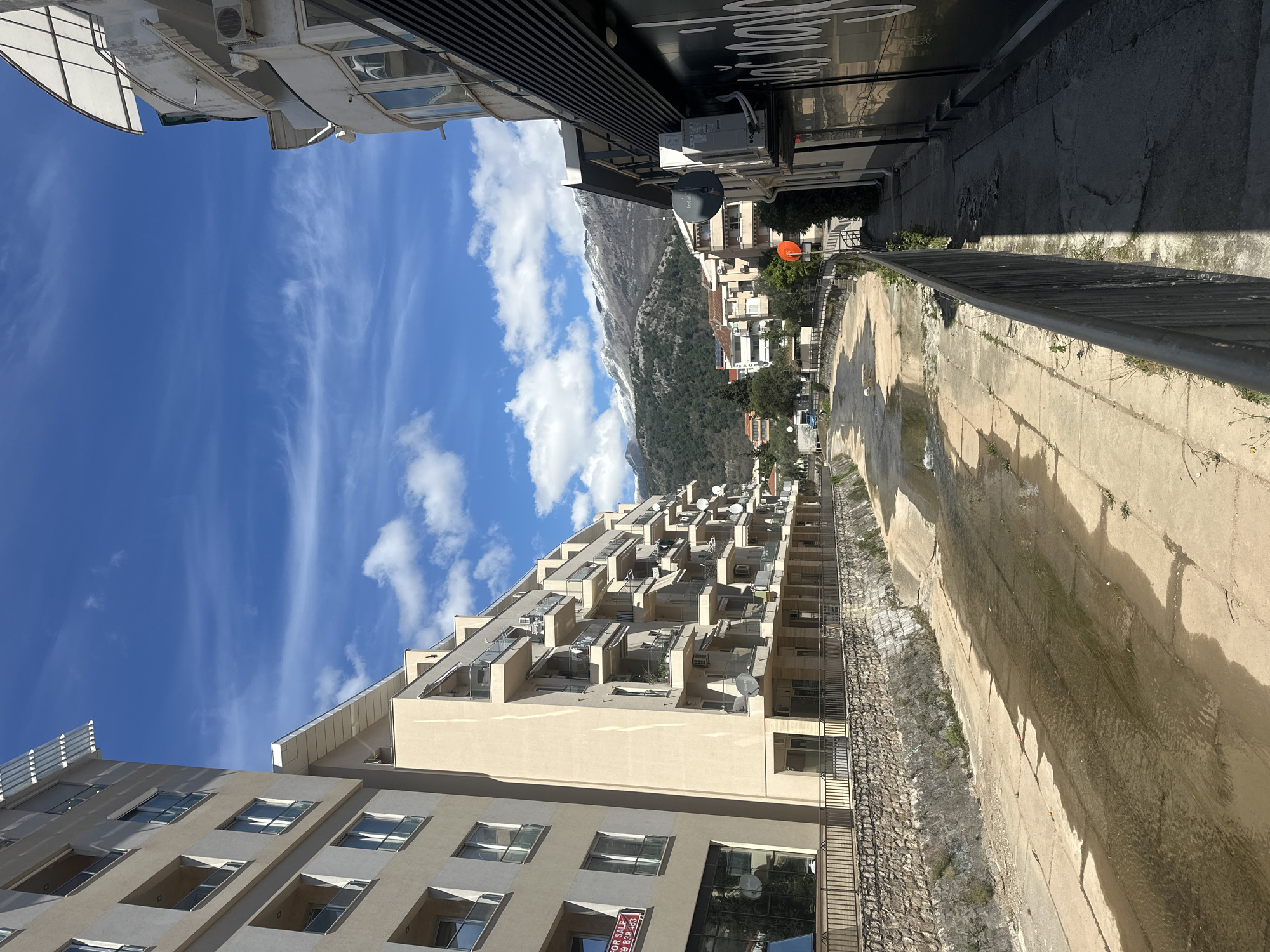 The image depicts a street scene in Budva, Montenegro, showcasing a drainage canal situated alongside a multi-story residential building. The building features numerous balconies, each equipped with satellite dishes. In the background, a mountain range with snow-capped peaks is visible. The sky is blue with scattered clouds. A narrow sidewalk runs parallel to the drainage canal. There are no people in the scene.