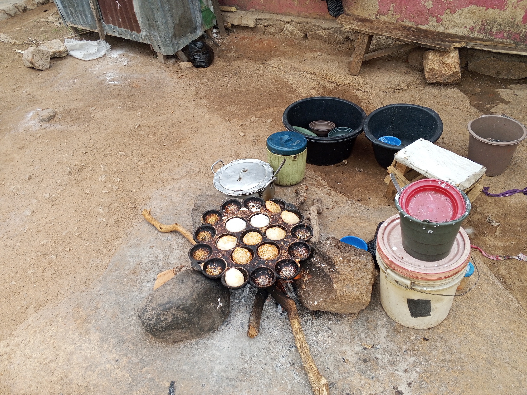 Outside in Gwong, Nigeria, a cast-iron pan sits on two rocks, heated by a small fire. The pan has multiple circular depressions, some filled with light-colored batter that is cooking, while others are empty and dark. To the right, a stacked arrangement includes a bucket filled with a pink substance, resting on another bucket. A white block rests on a wooden support. Two black basins and a brown bucket are positioned behind. In the background, a dilapidated wooden bench leans against a wall. A lean-to constructed of corrugated metal stands to the left. The ground is a mix of dirt and concrete.